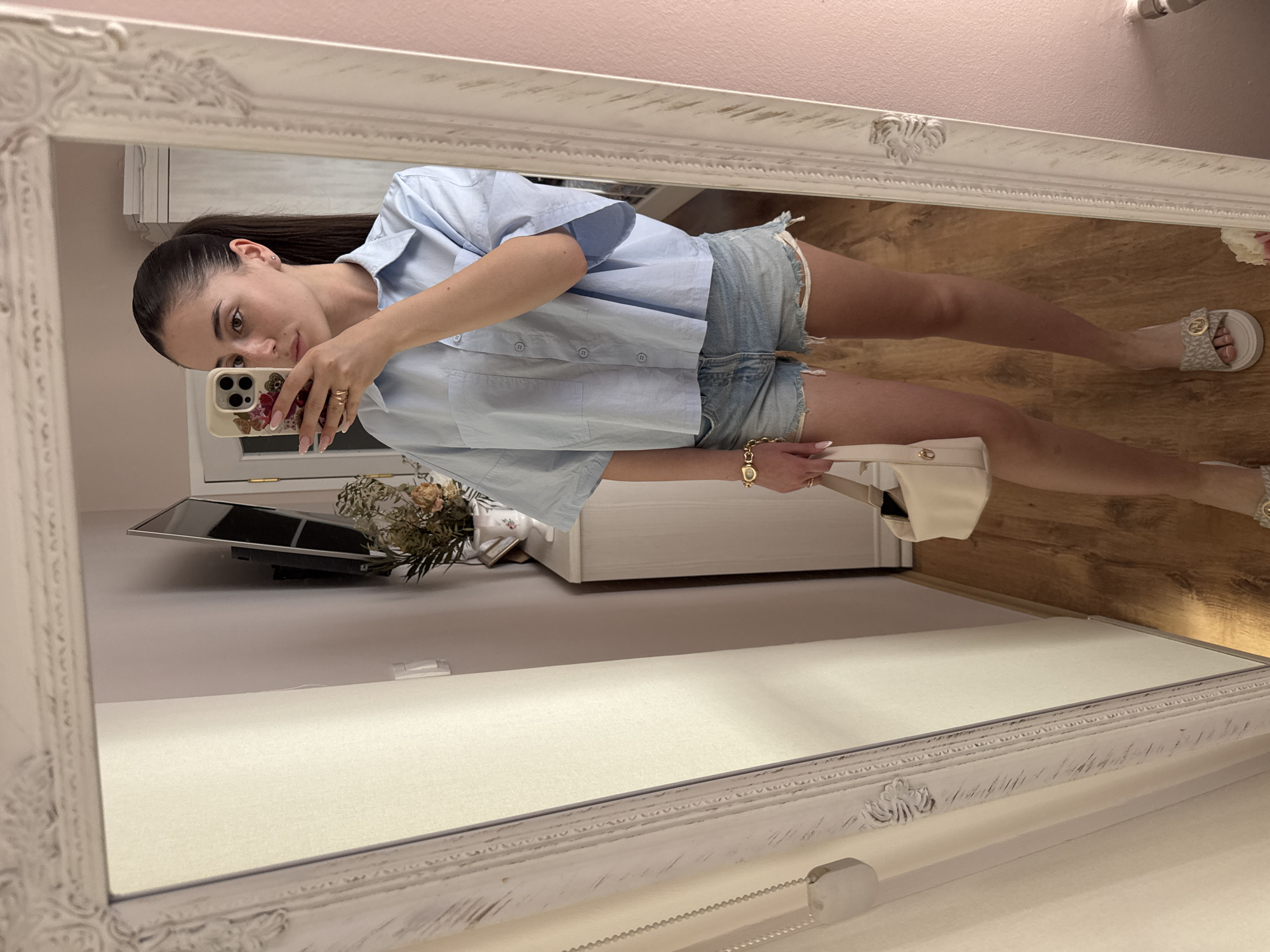A woman is taking a mirror selfie in a room. She is wearing a light blue short-sleeved button-up shirt, denim shorts, and slide sandals. She is holding a cream-colored handbag. A mirror with a distressed white frame is in the foreground. The room appears to be a bedroom or dressing area, with a wooden floor, a closet, and a television mounted on the wall. The lighting is bright and natural. The specific location in Targovishte, Bulgaria is not determinable from the image.