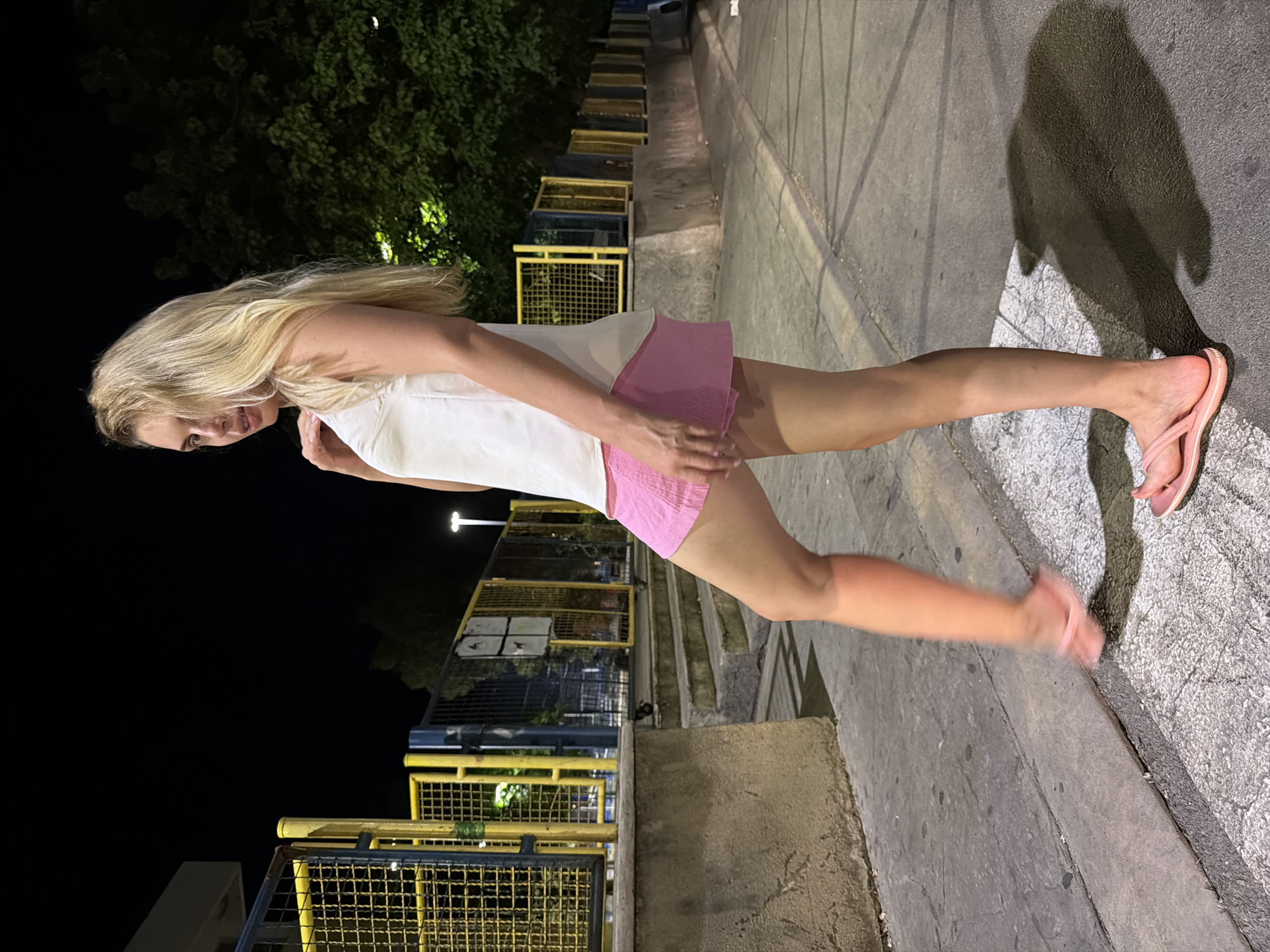 A young blonde woman stands on a paved area at night, with a dark background of trees and lights. She is wearing a white tank top and a pink skirt, and pink flip-flops. Her left leg is raised as if walking, and her right foot is visible at the bottom of the frame. She is looking over her right shoulder towards the camera with a slight smile. The scene appears to be in an urban environment, possibly a park or recreational area, with a yellow and blue fence in the background. The image has a cinematic quality with dramatic lighting and shadows.