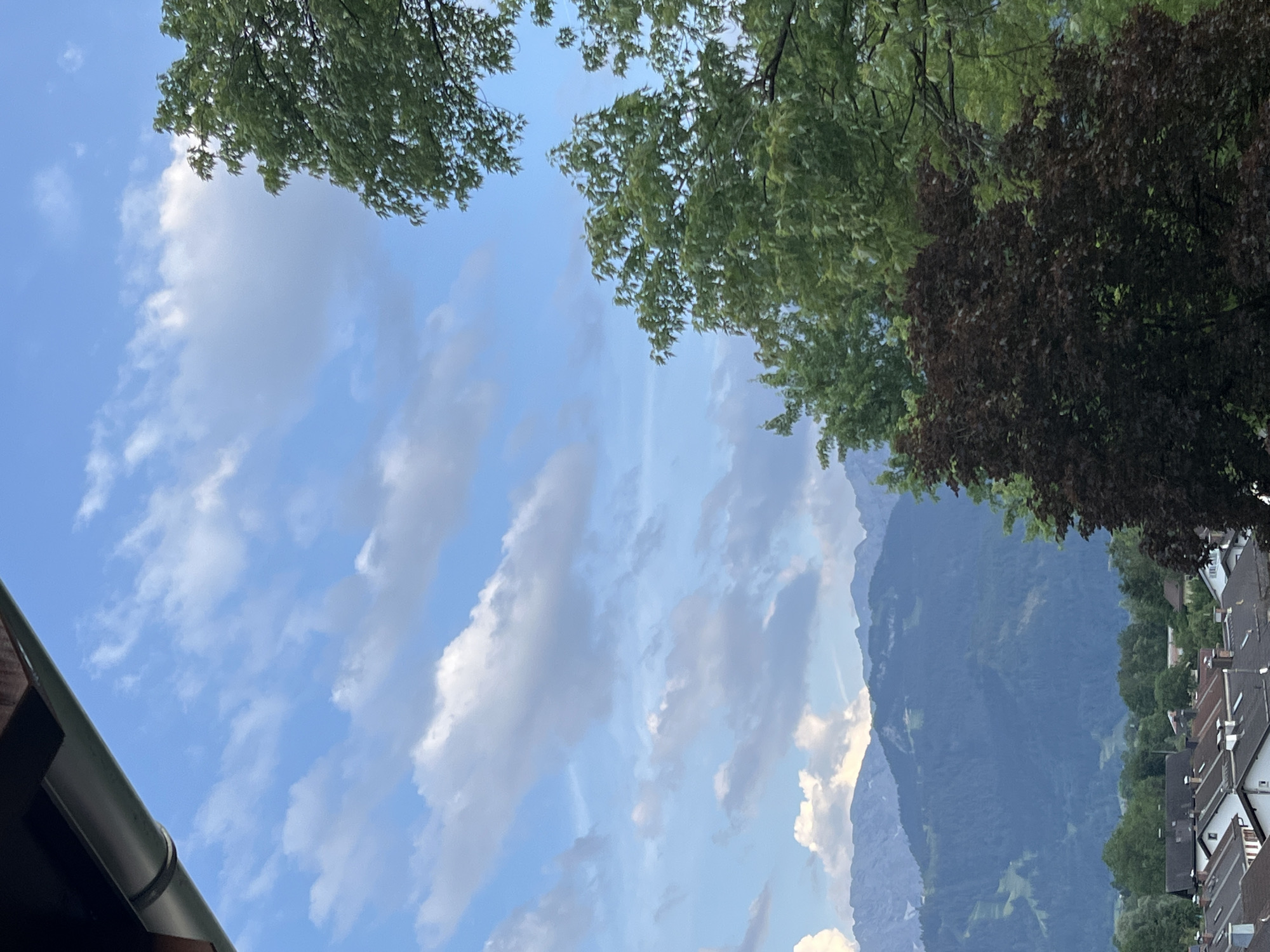 This media file captures an outdoor scene looking up at a bright blue sky dotted with fluffy white clouds. Lush green trees with dense foliage frame the top of the image, with a cluster of darker, reddish-brown leaves on the right. In the distance, a majestic mountain range is visible, its slopes covered in dense green forest. The lower right portion of the image shows a row of houses with distinctive pitched roofs, typical of a European village. A metal gutter runs along the bottom left edge, suggesting it is part of a building. The time of day appears to be daytime, with clear skies and sunlight illuminating the landscape. The weather seems pleasant and fair, conducive to outdoor activities. The scene evokes a sense of natural beauty and tranquility, with the imposing mountains juxtaposed against the cozy village.