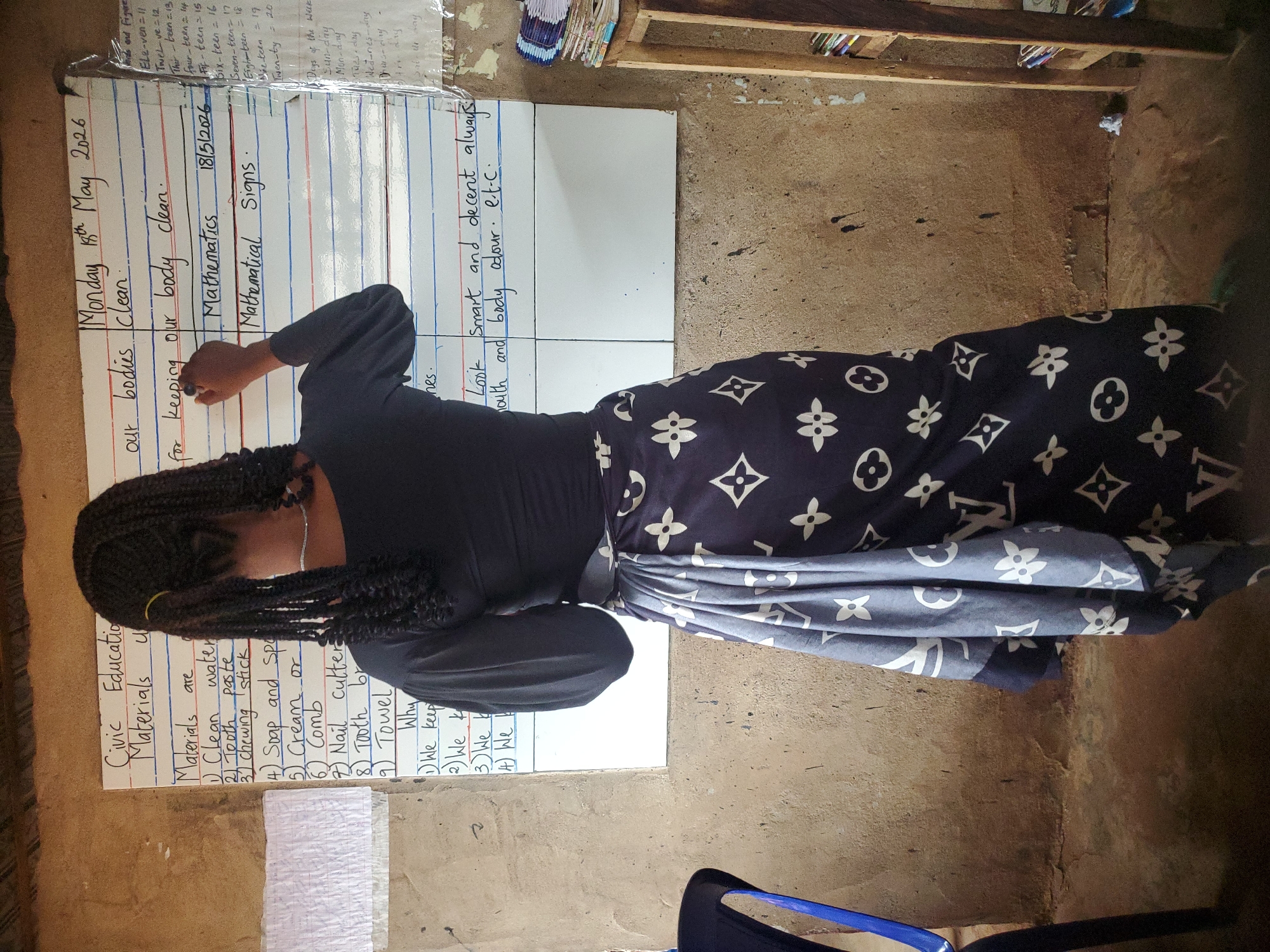 Here is a detailed description of the image:
**Scene:** The image appears to be taken in a classroom setting, based on the educational materials present. The environment looks like a rural area, the background is a rough, possibly unfinished wall. The location is specified as Ukwu-Abwa, Nigeria.
**People:** A woman stands in front of the whiteboard. She is dark-skinned and wears a black long-sleeved shirt and a patterned skirt/wrap. She has braided hair. She is in the process of writing on the whiteboard with a marker.
**Subjects & Objects:**
* **Whiteboards:** Three whiteboards are positioned side by side. One contains the date (Monday, 18th May 2026), the subject (Mathematics), and topics related to cleanliness. Another whiteboard has 'Look smart and decent always outh and body odour'
* **Educational Materials:** Other handwritten notes, possibly lesson plans or materials, are also visible on the whiteboards, covering subjects such as "Civic Education," "Materials are," and a list of items such as "Clean Water," "Toothpaste," "Chewing Stick," "Soap," "Cream," "Comb," "Nail cutter", and "Towel". Mathematical "Is and Figure's" lessons are also visible in the top left corner.
* **Classroom Furniture:** A blue plastic chair is visible in the foreground.
**Actions & Interactions:** The woman is writing on the whiteboard, suggesting that she is teaching or preparing a lesson.
**Overall Impression:** The scene shows a teacher in what appears to be a resource-constrained classroom setting, delivering a lesson on hygiene, cleanliness, and mathematical concepts. The rural location and the condition of the wall point to limited infrastructure.