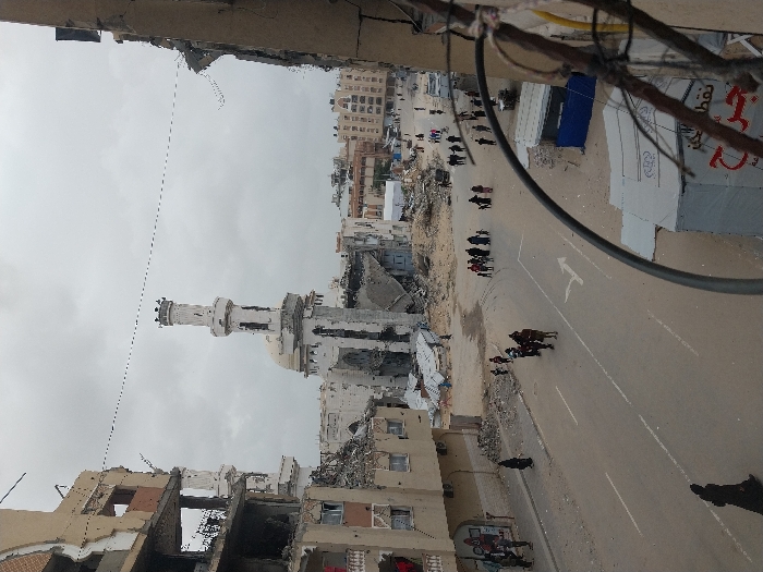 The image captures an elevated view of a street in Khan Yunis, Palestinian Territory, under an overcast sky. In the mid-ground, a large mosque with a prominent white minaret and dome exhibits significant structural damage, standing amidst rubble. To the left, a multi-story building displays extensive destruction, with entire sections of its facade missing, exposing internal structures and rebar. Another severely damaged building frames the right side of the foreground. The asphalt street below features a white turning arrow and double solid white lines. Approximately two dozen individuals, including men, women, and children, are observed walking on the street and adjacent dirt areas. One person leads a brown horse in the center of the street. Piles of concrete and debris are scattered along the road. In the right foreground, a white temporary tent structure with Arabic script and a smaller blue shelter are visible. Further damaged urban structures are discernible in the background.
