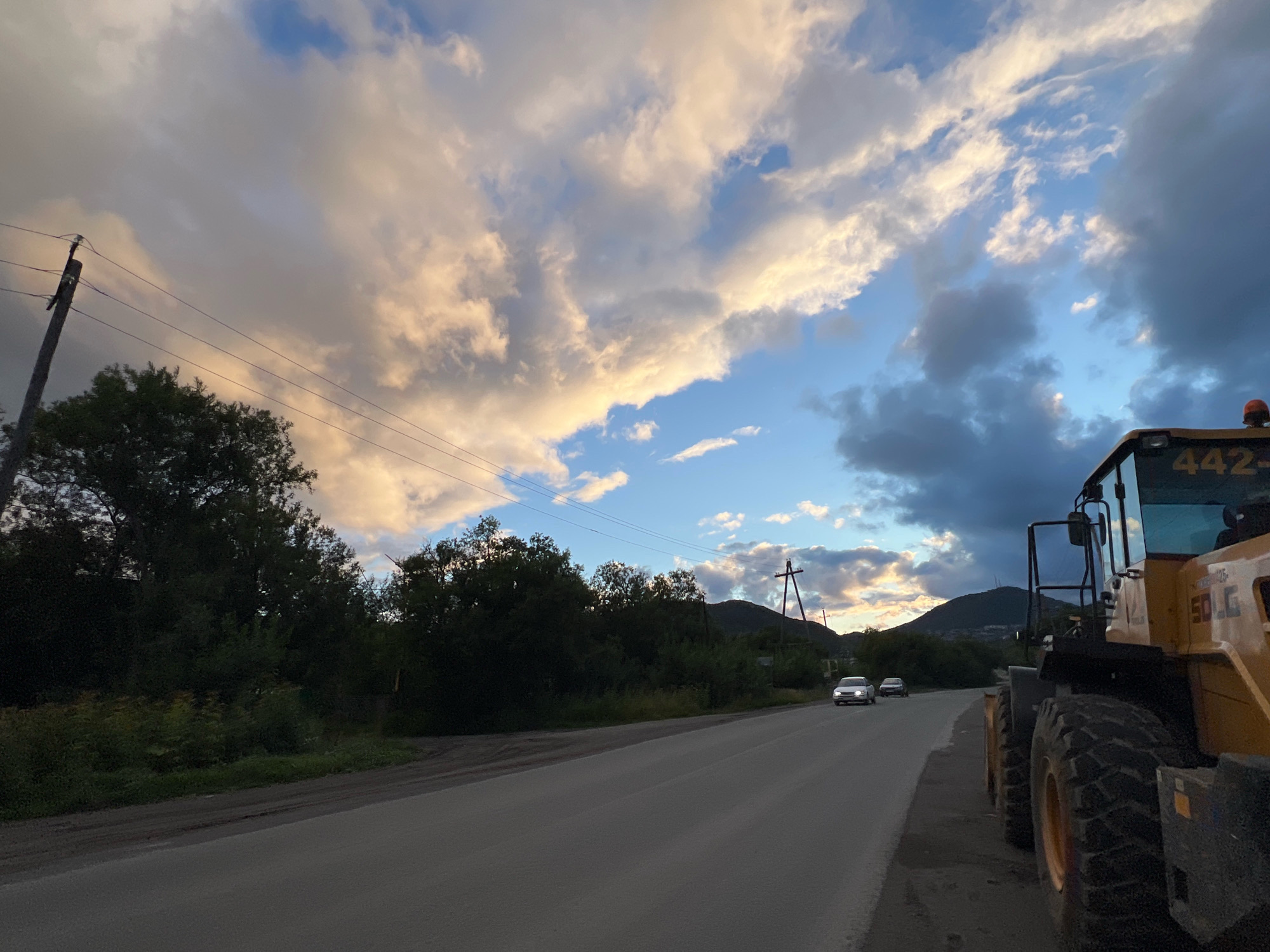 This is an outdoor, eye-level, medium shot captured in the city of Petropavlovsk-Kamchatsky, Russia. The scene depicts a roadside view under a dramatic sky filled with both light and dark clouds.
In the foreground, on the right side of the image, is the front section of a large yellow construction vehicle. Its massive tires and part of its cabin are visible. The vehicle has markings that include "50LG" and "442".
To the left of the construction vehicle, a paved road stretches into the distance. Two white cars are seen driving on the road. The left lane appears to be empty, while the right lane has traffic.
The roadside on the left is lined with dense green trees and foliage. A wooden utility pole with wires running across it stands prominently on the left side of the road.
In the background, rolling hills and mountains are visible, partially obscured by the trees and the receding road. The sky is a mixture of blue patches and large, textured clouds, some illuminated by sunlight, creating a sense of dusk or a transitioning weather condition. The overall impression is a rural or semi-rural setting on the outskirts of a city, likely during the late afternoon or early evening.