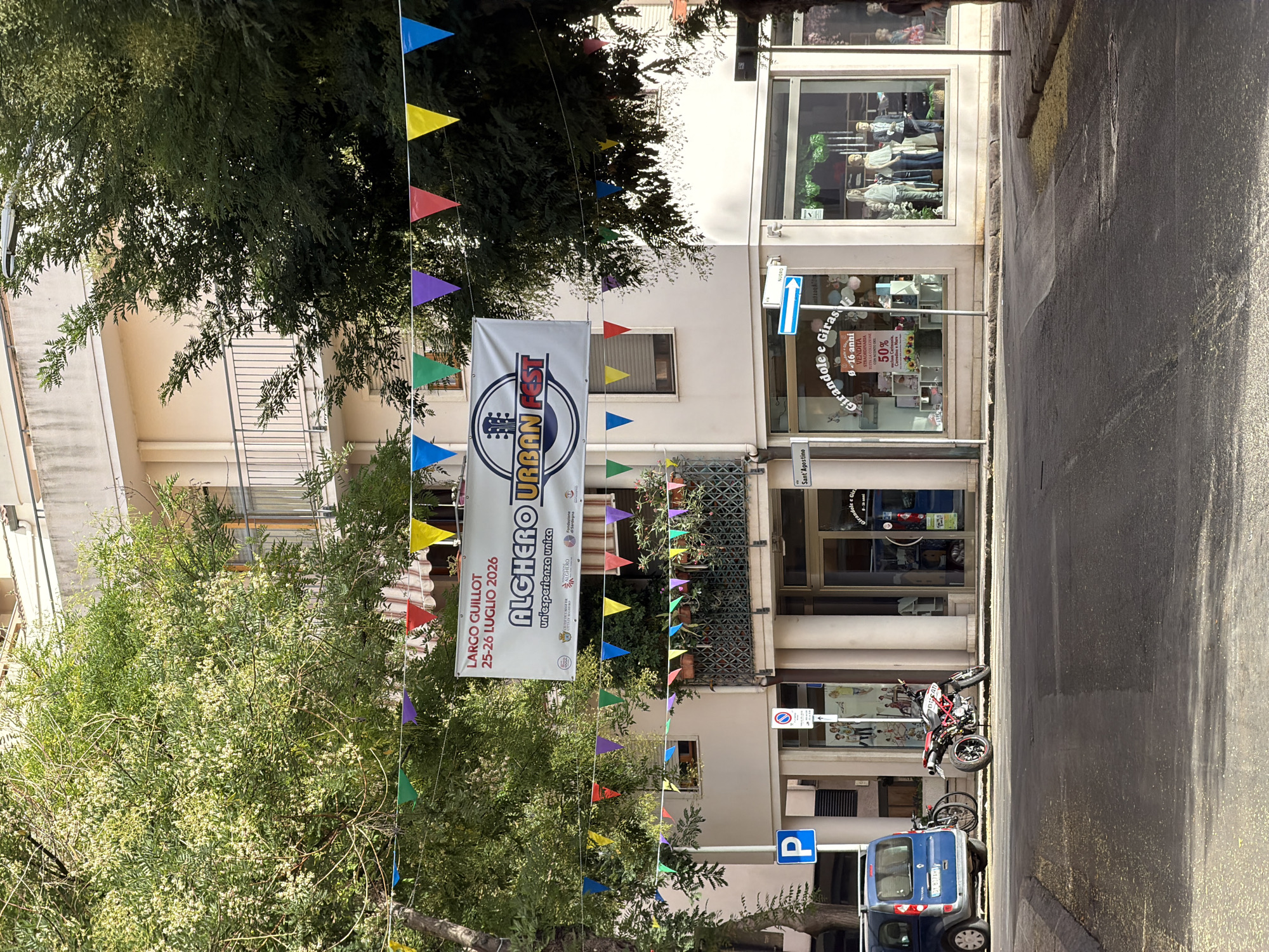 The image shows a street scene in Alghero, Italy, on a sunny day. A banner advertising "Alghero Urban Fest 25-26 Luglio 2026" is hung across the street, adorned with colorful triangular flags. Below the banner, a storefront features mannequins displaying clothing. A blue van is parked to the left, next to a bicycle. A red motorcycle is parked on the sidewalk in front of the storefront. Trees with green foliage are visible on either side of the street, casting shadows. The street itself is paved and appears to be a public area with buildings and shops lining one side. A blue "P" sign and a parking sign are also visible. The overall impression is a lively street scene, possibly set up for a local festival.