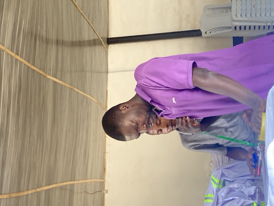 In an indoor facility in Agege, Nigeria, two adult males are observed. The prominent figure in the foreground, a man with dark hair and a short beard, wears a purple polo shirt featuring a white feline logo on the left chest. He is leaning forward, directing his gaze and hands toward a white fabric spread on a surface. Adjacent to his hands are a yellow sponge-like object, a red-capped pen, and a blue tool. Behind and to the left, a second man in a grey t-shirt, with a green measuring tape draped around his neck, also looks down intently at the work surface; his right hand rests near his chin. To the left of the men, numerous stacks of neatly folded white garments, some exhibiting yellow-green reflective bands, are visible. On the right side of the frame, a stack of light-colored plastic chairs is present. The environment consists of light-colored walls and a ceiling composed of corrugated material supported by wooden beams. A black vertical support or pipe is situated on the right, and an exposed light fixture hangs from the ceiling on the left.