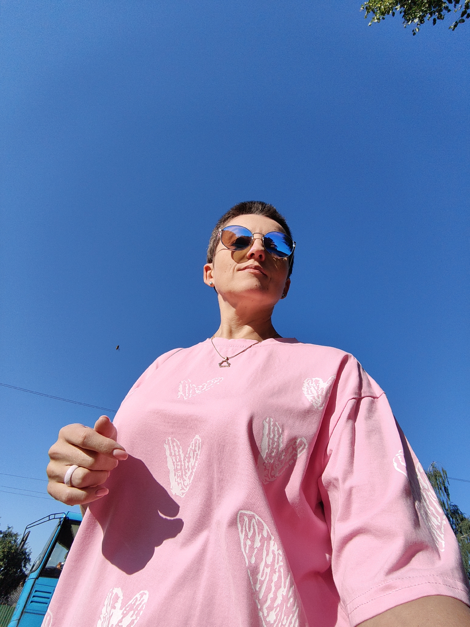 A low-angle, full-body, outdoor photograph captured on a bright, sunny day shows a woman standing against a clear blue sky. The woman, who appears to be an adult, is wearing a bright pink t-shirt with white heart patterns and reflective sunglasses. She is looking upwards and to her right, with her left arm bent and her hand near her chest. A light-colored ring is visible on her left ring finger. A small bird is visible in the sky to the left of the woman. In the background, a portion of a blue vehicle and some green foliage are visible. The setting is implied to be the city of Fanipal, Belarus.