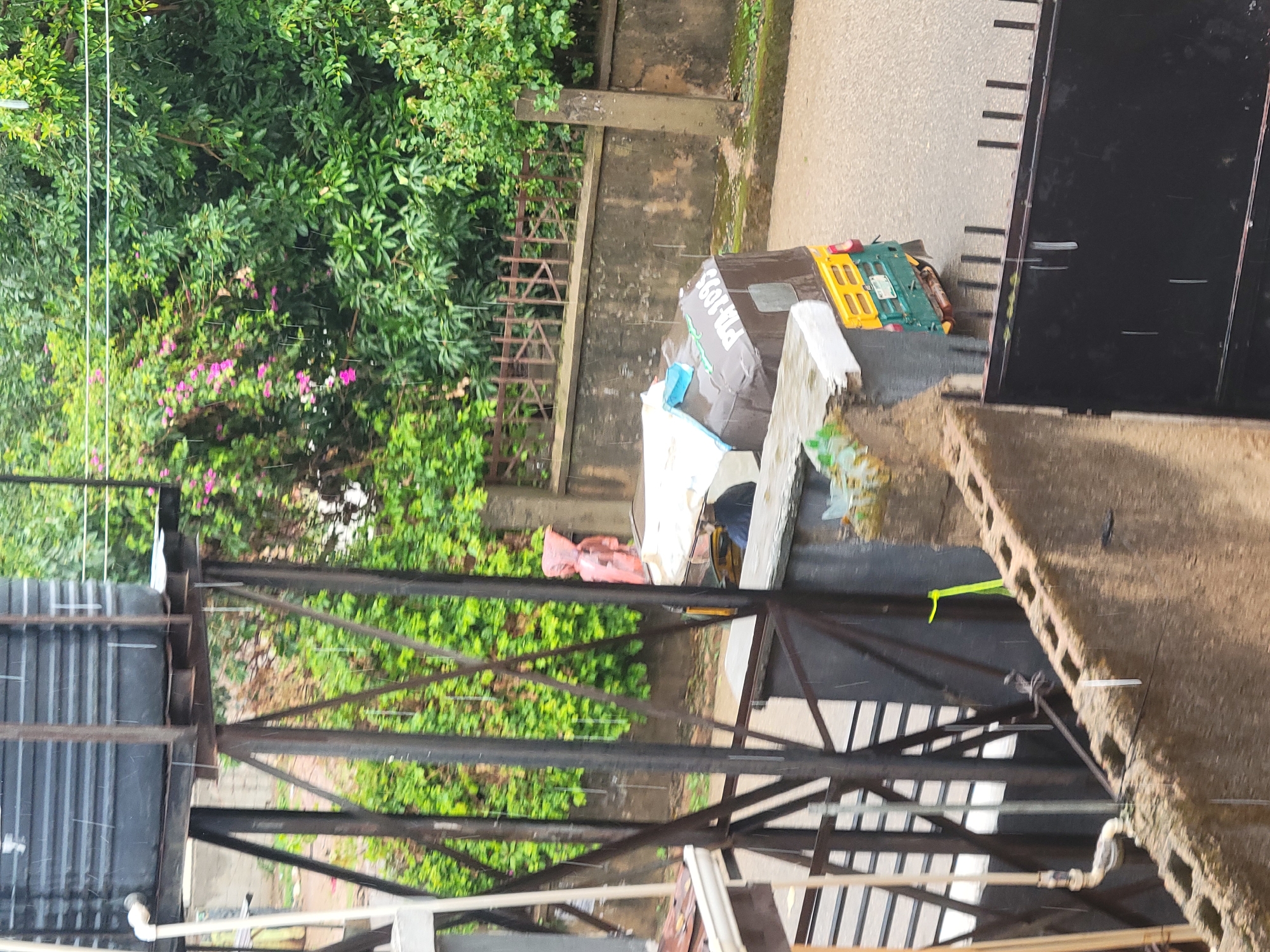 A close-up, medium shot captures a rain-soaked scene from an elevated perspective, looking down and across a street. In the foreground, a rough, concrete block wall with visible rebar protrudes into the frame, partially obscuring a dark, metal gate with vertical bars. To the left, a substantial black water tank is elevated on a rusty metal framework, with a white pipe extending from its side. Heavy rain is evident, with visible streaks falling through the air and wetting surfaces.
The middle ground reveals a street with a wet asphalt surface. A three-wheeled auto-rickshaw, known locally as an "okada" or "keke," is parked on the right side of the street. It is primarily brown with a yellow roof and a green and yellow lower body. The rickshaw's license plate is partially visible, reading "PTA-1095." A person, possibly the driver, is seated inside the rickshaw, wearing dark clothing and a hat. Another figure, wearing a bright pink garment, stands near the rickshaw.
The background is dominated by lush green foliage and trees, with hints of pink flowers. A rustic wooden fence runs along a wall that separates the street from the vegetation. The overall impression is of a residential or semi-urban area in Jos, Nigeria, experiencing a downpour.