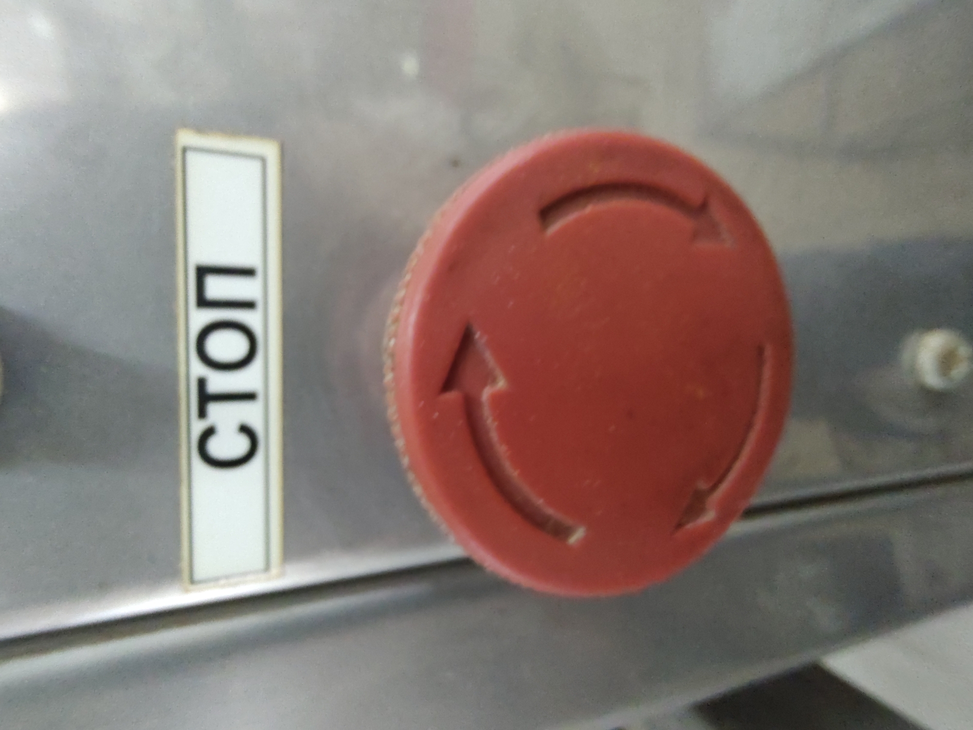 The image shows a close-up of a red emergency stop button on a metallic surface. Above the button, a rectangular label with a white background and black text reads "СТОП" which is the Russian word for "STOP". The button itself is circular with a textured edge and features three indented arrow shapes pointing in a clockwise direction around its circumference. The metallic surface appears to be part of a machine or industrial equipment, with a brushed texture. The lighting is overhead and casts a slight shadow. There are no people or animals visible in the frame. The image does not provide any information about the city of Tervel, Bulgaria.