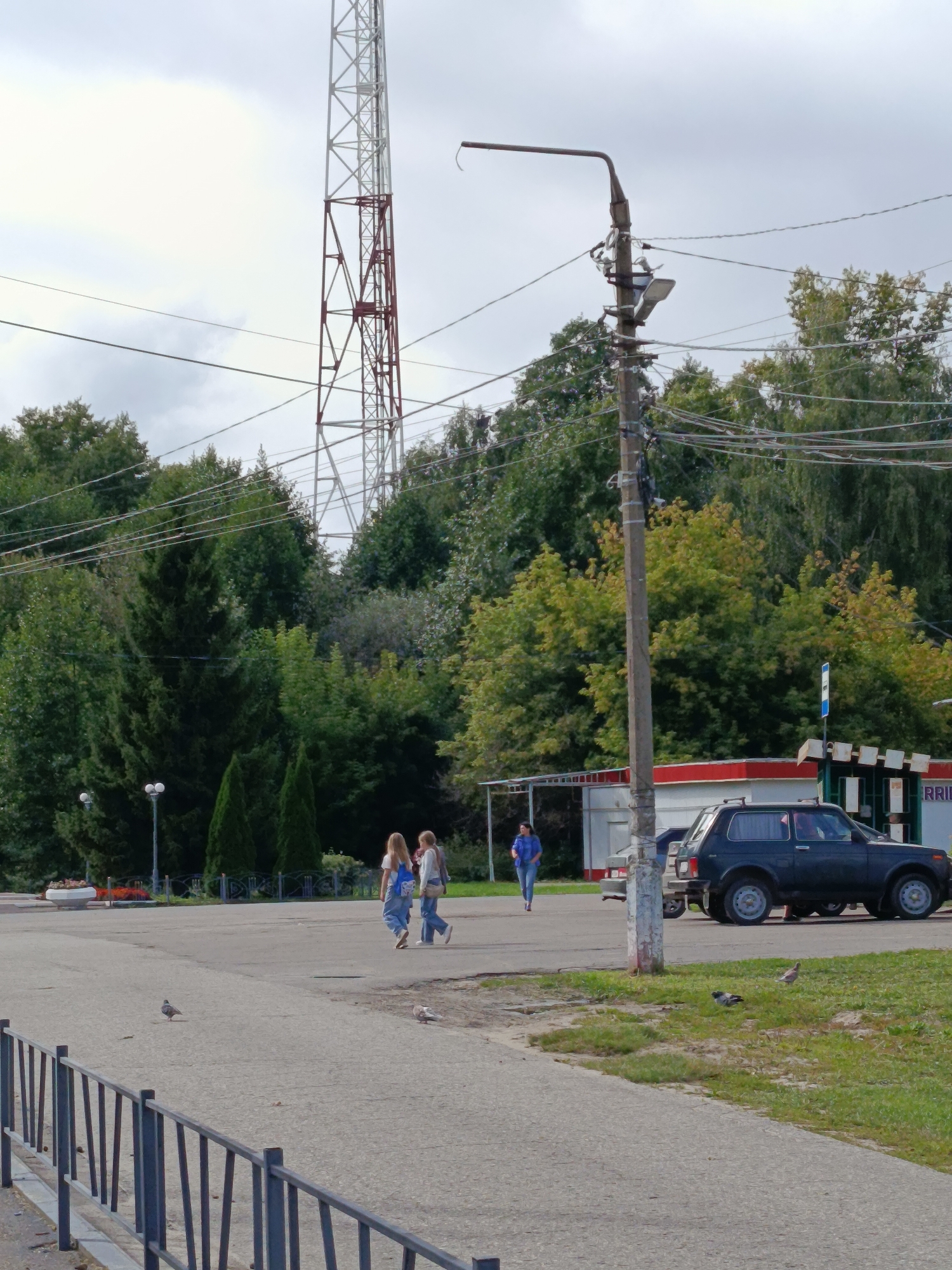 The image depicts an outdoor scene in Verеya, Russia, under an overcast sky. In the background, a tall, red and white lattice tower dominates the skyline, with numerous power lines crisscrossing the view. A concrete lamppost with electrical equipment stands in the mid-ground. To the right, a dark-colored SUV is parked near a covered structure that appears to be a bus stop or a small kiosk with a red roof. A woman in a blue jacket and jeans is walking towards the left of the frame. In the mid-ground, two young women in casual clothing are walking away from the camera, one carrying a blue backpack. A paved pathway extends into the foreground, with a metal fence bordering the lower left. Two pigeons are visible on the pavement. Trees with green and yellow foliage form a dense backdrop behind the structures and pathway.