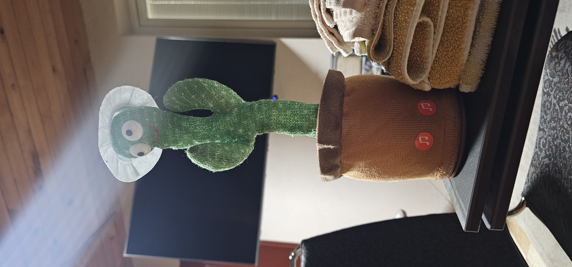 A plush cactus toy wearing a white hat is positioned on a shelf. The cactus is green and appears to be inside a brown pot with red musical note symbols. The shelf also holds folded beige and tan towels. In the background is a television and part of a wooden wall. The location of the image is unknown.
