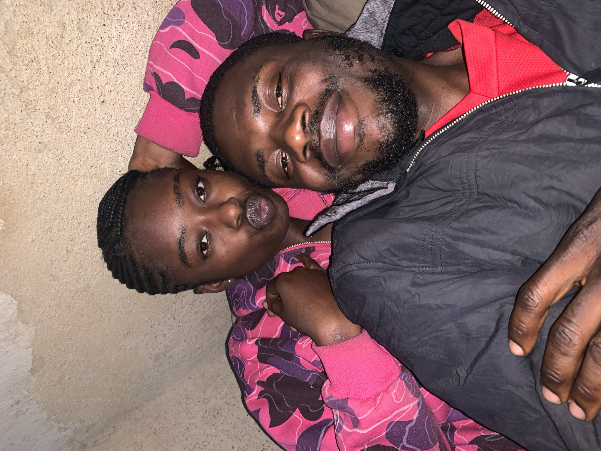 A close-up, selfie-style photograph captures two individuals in what appears to be an outdoor, dimly lit environment, possibly a wall or alcove. The person in the foreground is a man with dark skin, a beard, and a goatee. He is wearing a black jacket zipped open over a red shirt. His eyes are closed, and he has a slight smile. To his left, and slightly behind him, is a young woman with dark skin and braided hair. She is wearing a pink and purple camouflage-patterned hooded jacket. She is looking towards the camera with her lips pursed in a kissing gesture. Her left arm is resting on the man's shoulder, and her right hand is touching her face. The background is a textured, light-colored surface, possibly a concrete wall, with some visible discoloration and imperfections. The overall lighting is warm and suggests an evening or nighttime setting. The photograph is taken from a slightly low angle, looking up at the subjects.