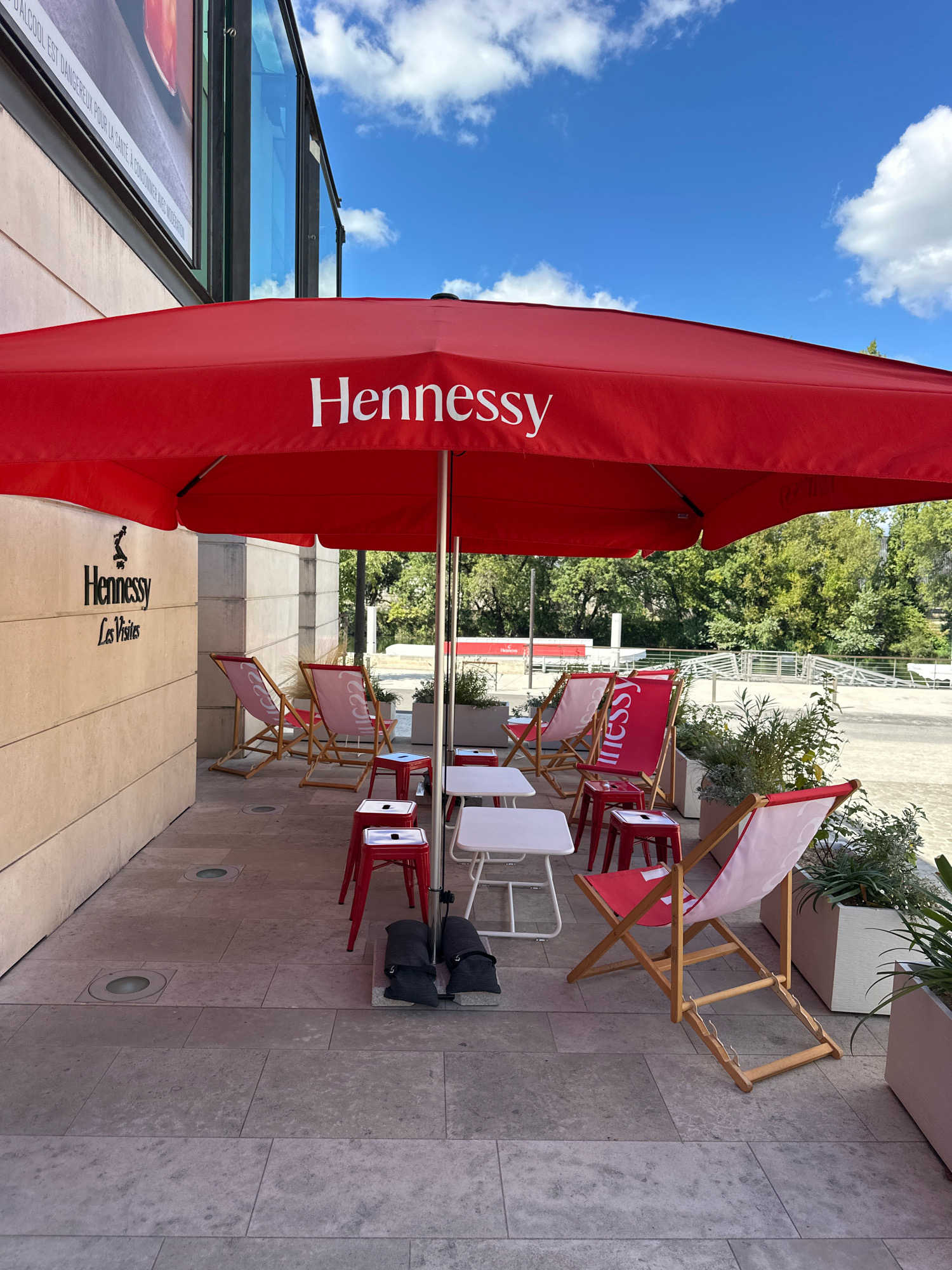 The image shows an outdoor seating area in front of a building with a Hennessy advertisement and branding. A large red umbrella with the "Hennessy" logo prominently displayed shades a seating area with several deck chairs and small tables. The chairs are red and white, and some have the Hennessy logo on them. There are also small red stools and white square tables. The building facade is made of stone, and there is a planter with greenery to the right of the seating area. In the background, trees and a blue sky with clouds are visible. The scene appears to be set in Cognac, France, given the prominent Hennessy branding, which is a renowned cognac producer based in the city. The area is paved with stone tiles, and there are recessed lights in the pavement.
