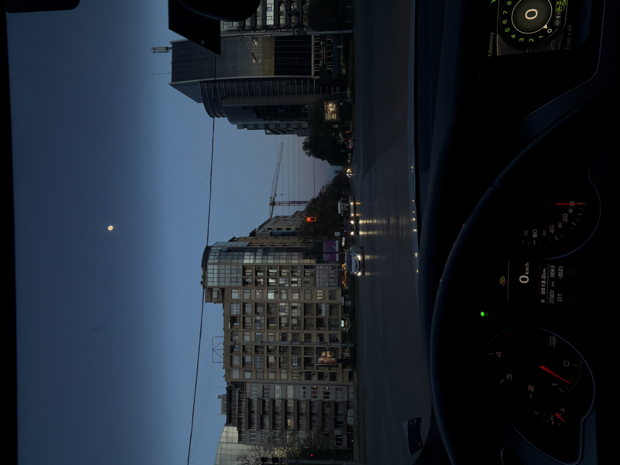 The image captures a twilight or nighttime urban street scene from the perspective of a car's interior, likely in Sofia, Bulgaria. The sky is a deep blue, with a bright moon visible on the left side.
In the middle and background, various multi-story city buildings dominate the landscape. A prominent building on the left features numerous windows and a modern facade. Another building in the background displays a banner, partially readable as "HOME CAPITAL". A tall construction crane stands out against the sky in the central part of the image. The street appears wet, reflecting the headlights of other vehicles and streetlights, suggesting recent rain. A red traffic light is visible in the distance.
On the right side, the interior of the car is visible, including the steering wheel and parts of the dashboard and infotainment system. The dashboard instruments show the vehicle is stationary, with the speedometer at "0 km/h." The odometer reads "273837 km," with trip meters displaying "9518.0 km" and "1656.6." The outside temperature is "+20.0°C," and the gear is indicated as "D1." The car's infotainment screen displays a numeric keypad (0-8) with options like "Memory" and "End call," and an "A-Z" symbol. The scene conveys a calm, urban evening ambiance.
