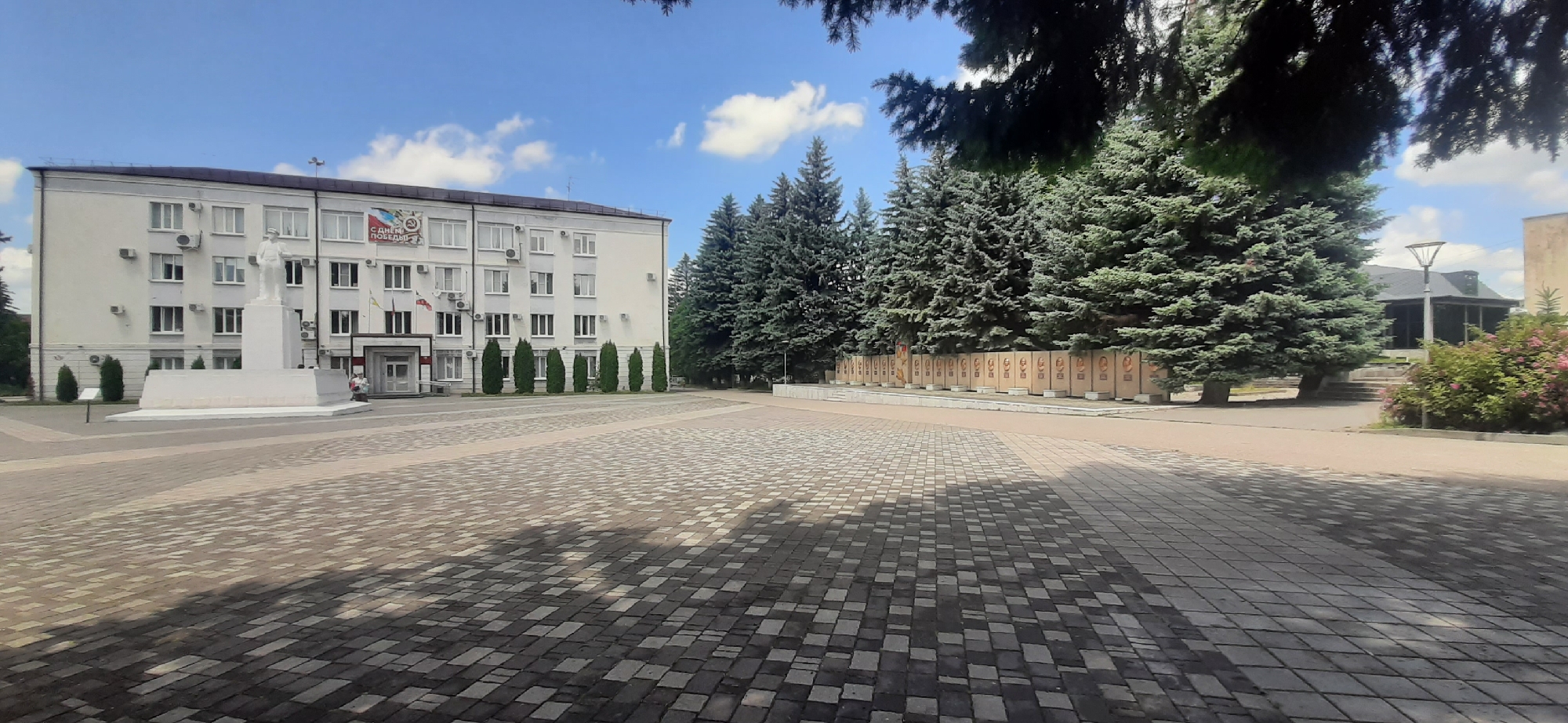 The image shows a wide paved plaza in front of a white, multi-story building. The building has numerous windows and is decorated with flags and a banner that reads "С ДНЕМ ПОБЕДЫ!" (Happy Victory Day!). A white statue of a man stands in front of the building on a raised platform. Several tall evergreen trees are planted in a row behind the building, and further to the right, a wall with many framed portraits is visible, partially obscured by more trees. A path leads away from the plaza towards a building with a dark roof and a lamppost. The plaza is paved with interlocking bricks in a patterned arrangement, and shadows from nearby trees are cast across it. The sky is blue with scattered white clouds. The overall scene appears to be a public space, possibly a government building or memorial site, in Yessentukskaya, Russia.