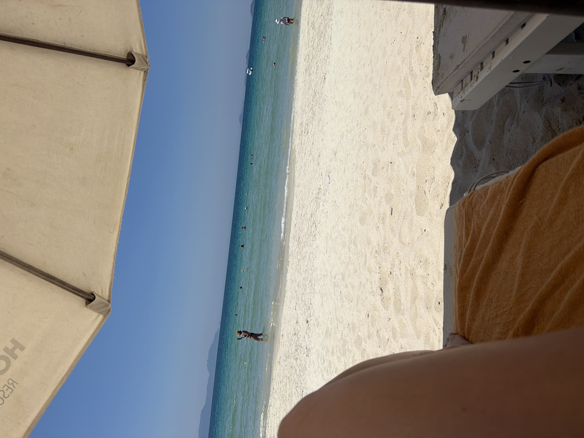 The image presents a first-person perspective of a beach scene, identified as being in Dong Ninh Hoa Ward, Vietnam. The upper quarter of the frame shows the underside of a beige beach umbrella or canopy, with partially reversed text "HOTEL RESORT" visible on its left side. Below the umbrella, a clear blue sky extends down to a horizon marked by a hazy line of distant mountains or landmass.
The middle ground is dominated by calm, turquoise-blue ocean water. In the water, several individuals are present. On the left side of the water, a person in dark swimwear stands in the shallow area, holding a smartphone at arm's length, seemingly capturing an image or video. Further out in the water, multiple other people are visible wading or swimming, along with a few small buoys. On the right side of the water, closer to the shore, a small child wearing a white hat and red attire stands in the shallow water. A small white boat is visible further out in the sea on the right.
The foreground consists of light-colored, fine-grained sand. In the extreme lower-left, a tanned human leg is visible diagonally. An orange towel is draped over a beige lounge chair, presumably where the viewer is positioned. Shadows, cast by an umbrella and another lounge chair, are visible on the sand. Part of a white wooden lounge chair frame with a beige cushion is also visible on the lower-right side of the frame.