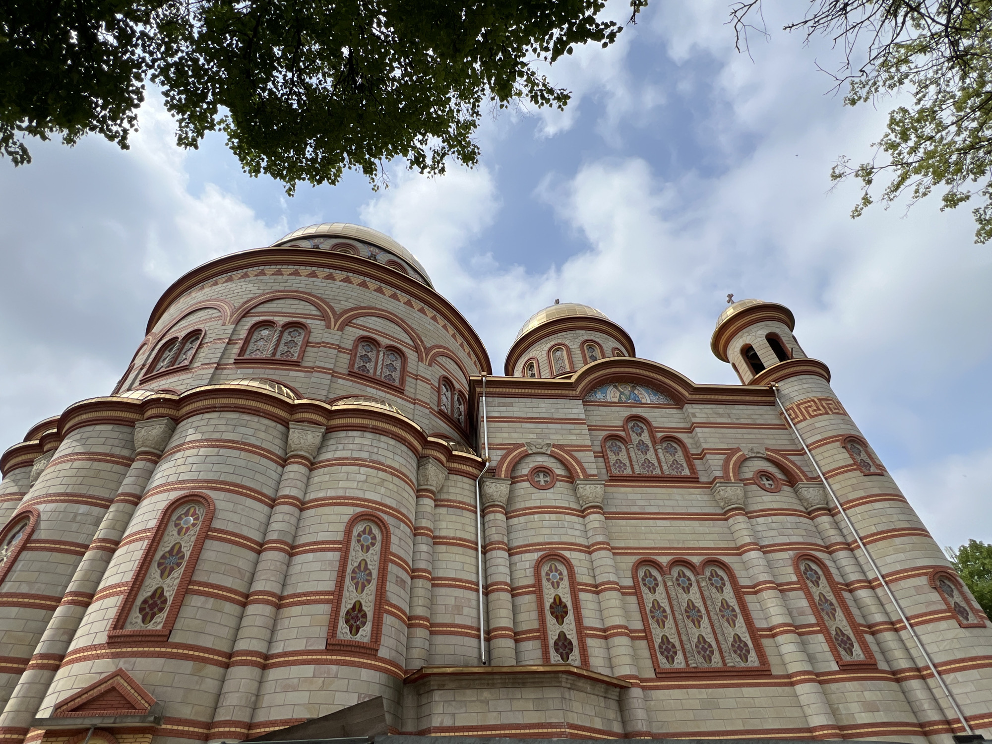 This low-angle, upward shot depicts a grand Eastern Orthodox church or cathedral, showcasing its ornate architecture against a partly cloudy sky. The magnificent building features a facade constructed from light-colored stone or brick, accentuated by horizontal bands of red brick and thin golden outlines. Multiple prominent golden domes crown the structure, with a larger one on the left and smaller ones towards the right, including one atop a small bell tower which is surmounted by a cross.
The walls are adorned with numerous arched windows, many of which contain intricate mosaic-like patterns or stained glass in floral and geometric designs, incorporating colors such as red, blue, and gold. Decorative columns and sculpted elements further enhance the building's elaborate appearance. Lush green tree foliage partially frames the top of the image, suggesting a natural outdoor setting.
The photograph was taken during daytime, with a bright yet diffused light illuminating the building under a sky filled with a mix of white clouds and patches of blue. No people are visible, and there is no specific activity or event captured in the frame. The architectural style, particularly the golden domes and detailed brickwork, is characteristic of religious buildings in Eastern Europe, consistent with the location context of Ternopil, Ukraine. No visible text is discernible on the building.