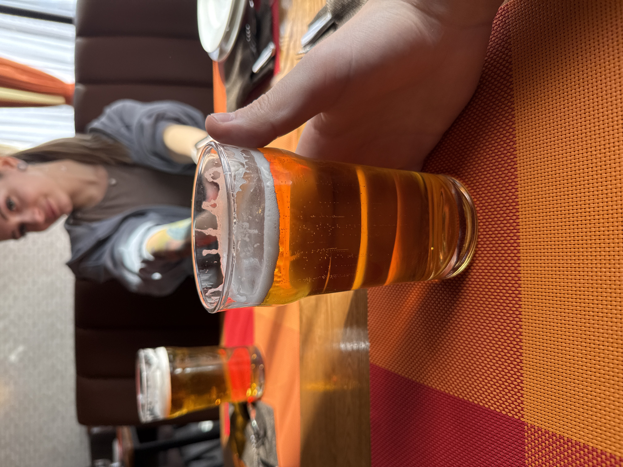 A photograph taken indoors in what appears to be a restaurant setting. A partially-filled glass of beer is centrally positioned, held by a person's hand. The background includes another individual seated at the table, partially visible, and dining utensils. The scene is set on a checkered placemat in shades of orange and red. Based on the provided information, the location is Moscow, Russia.