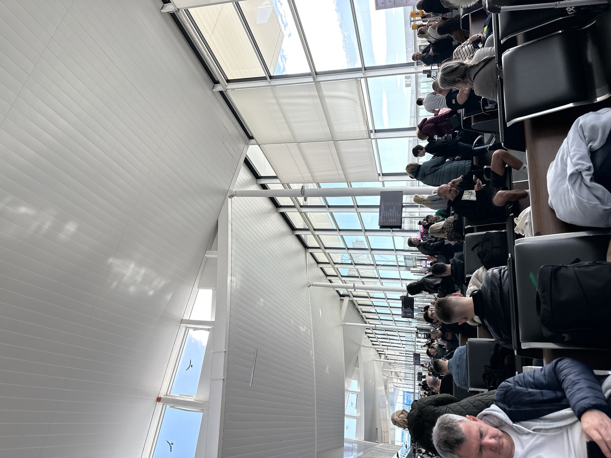 The image shows the interior of Wrocław Airport in Poland. Passengers are sitting in rows of chairs, presumably waiting for a flight. The airport architecture features a high ceiling with a grid-like structure of windows, allowing ample natural light to enter. Some windows have partially closed blinds. The walls are white and have a linear, panel-like texture. People are dressed in casual clothing, and some have luggage beside them. The scene conveys a sense of travel and anticipation.