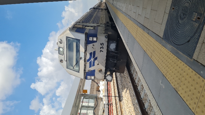 A daylight vertical photograph depicts a stationary Israel Railways locomotive, identified by the number "735" and the company's silver-grey and blue livery, at a train station platform in Rishon LeZion, Israel. The front of the locomotive, featuring its operational lights and insignia, is positioned on the track directly adjacent to the concrete platform. The platform includes a yellow tactile warning strip with raised dots along its edge and a dark, circular manhole cover visible in the foreground. To the left of the train, multiple railway tracks with gravel ballast are present. A modern white canopy structure extends over a portion of the platform in the upper left. Further in the background, an area with construction materials and red safety barriers is visible next to the tracks, with multi-story residential buildings discernible in the distance. The sky above is blue with scattered white clouds. No individuals are visible within the frame.