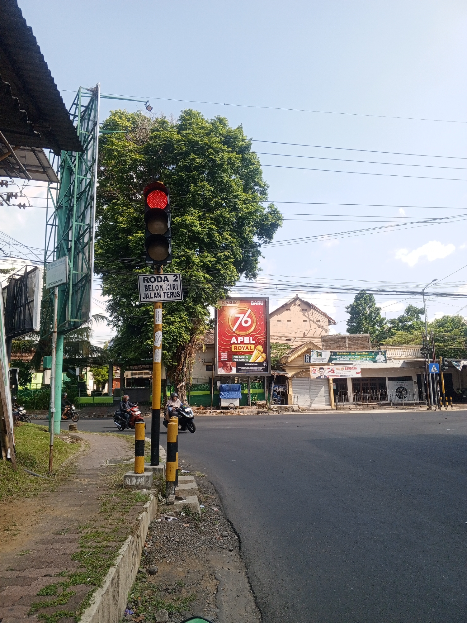 The image shows a street intersection in Jember, Indonesia, on a clear, sunny day. A traffic light is prominently displayed, showing a red signal. A sign below the traffic light reads "RODA 2 BELOK KIRI JALAN TERUS" which translates to "2 WHEELS TURN LEFT CONTINUE STRAIGHT." Several motorcycles are present on the road. In the background, there are buildings, trees, and utility poles with overhead wires. A large billboard advertising "76 APEL ROYAL" is visible on the right side of the intersection. A pedestrian crossing is marked on the road. The overall scene depicts a typical urban street environment.