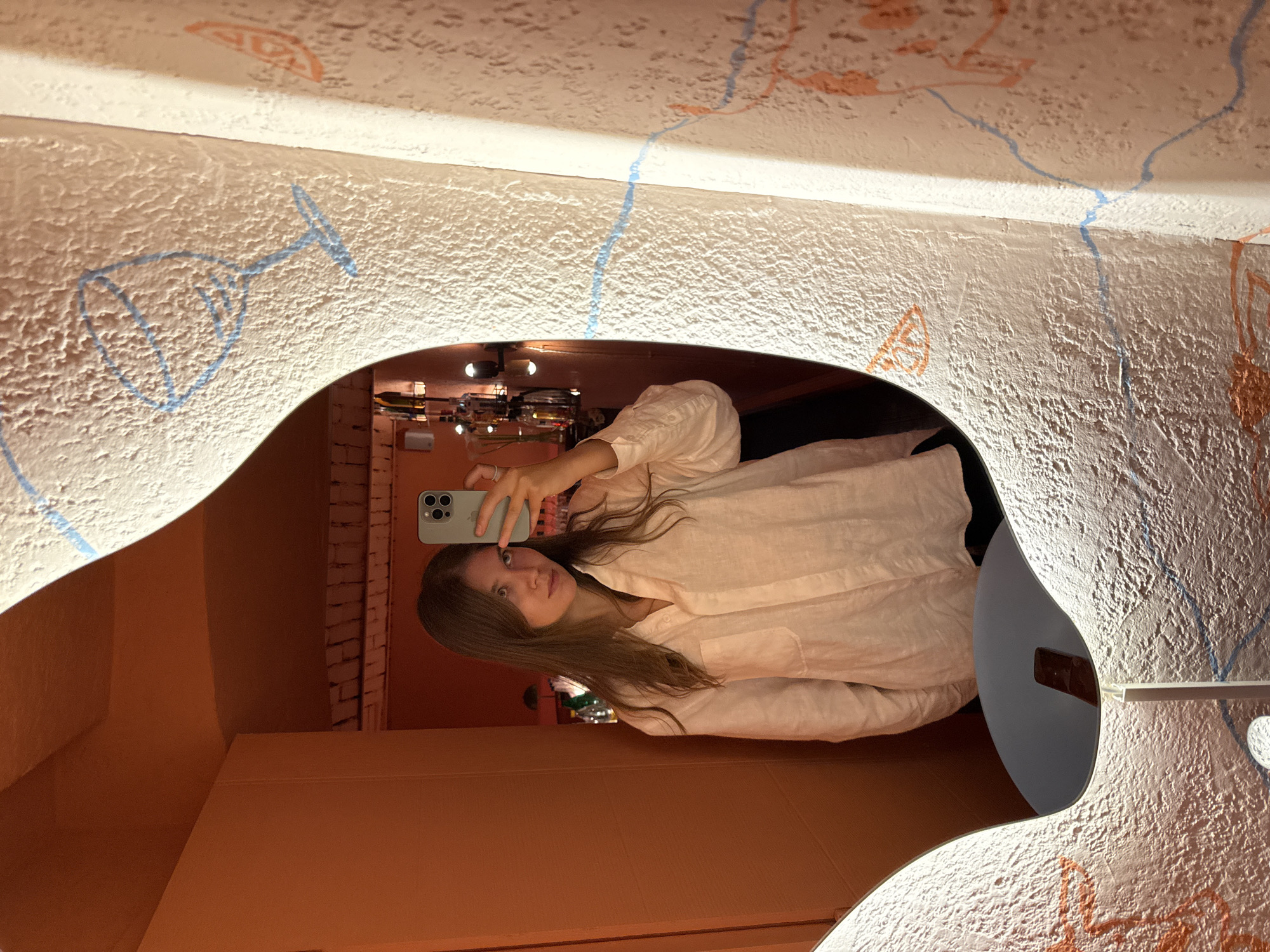 A young adult female is captured in a mirror selfie within an establishment located in Vracar Urban Municipality, Serbia. The woman, with long brown hair, wears a light-colored, loose-fitting collared shirt over dark clothing. She holds a light-green smartphone with three camera lenses in her right hand, positioned to capture her reflection; a silver ring is visible on her right ring finger. Her gaze is directed towards the camera.
The mirror itself is irregularly shaped, with an illuminated border, and reflects an interior space painted a uniform warm orange-red hue. Visible in the reflection are a plain orange wall to the left and, through an arched opening on the right, a dimly lit bar area with various bottles on shelves and warm-toned light fixtures. The ceiling in the reflected area appears to have exposed wooden beams or similar architectural detail.
The wall surrounding the mirror is textured with a rough, stucco-like finish and is adorned with various hand-drawn-style illustrations in blue and orange. These include a blue outline of a wine glass and abstract wavy lines, alongside orange outlines of fruit slices and other undefined shapes. A small, circular light fixture on a slender stand is visible at the bottom edge of the mirror, with a dark, rectangular object (possibly a remote control) resting on the surface directly below the mirror's reflective surface.