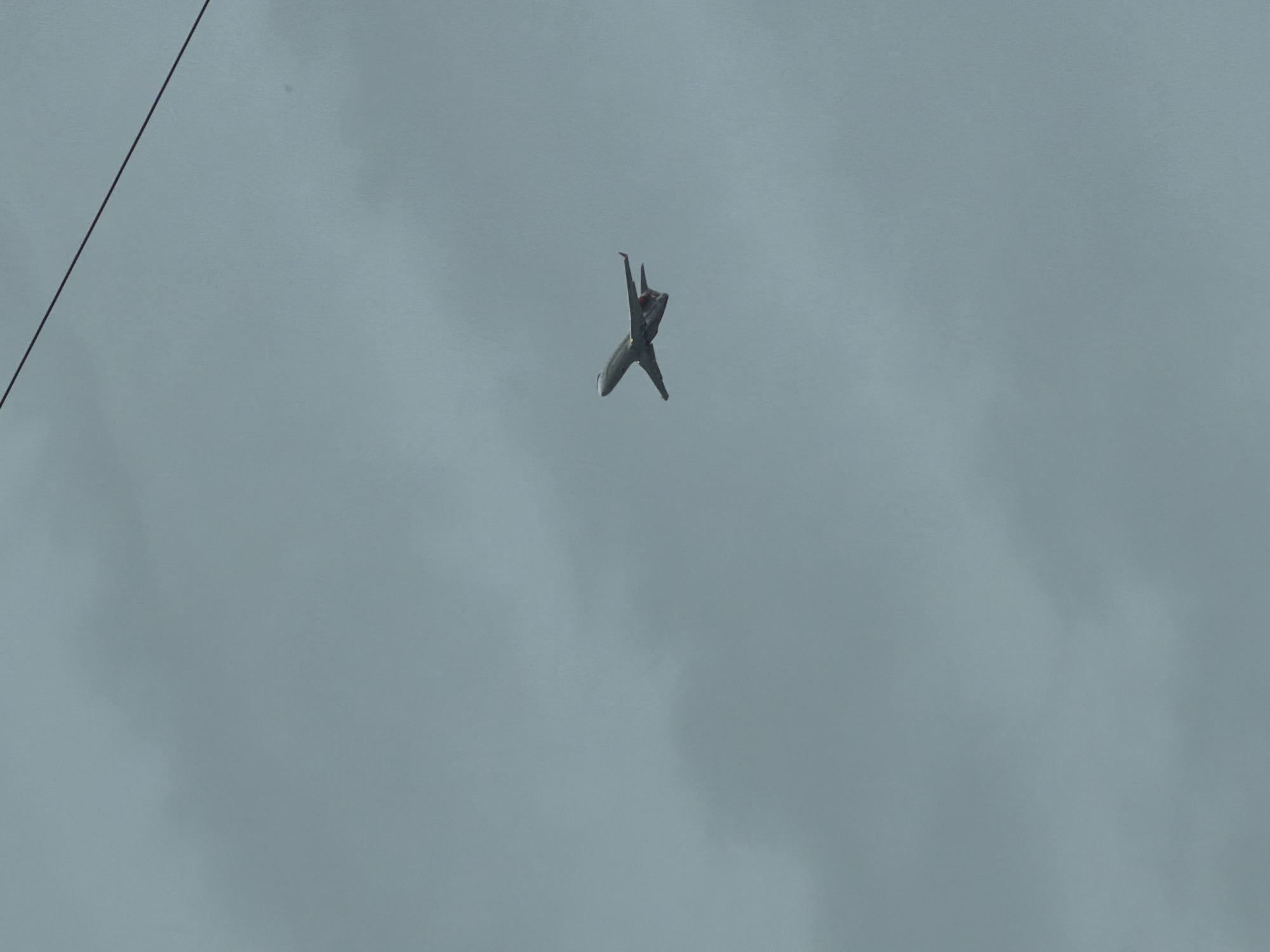 The image depicts a Sukhoi Su-30SM fighter jet in flight against an overcast sky. The aircraft is oriented with its nose pointing slightly downward and the wings angled back, suggesting it is performing a maneuver. A thin, dark cable is visible in the upper left corner of the frame. The location is likely near Perm, Russia, given the description.