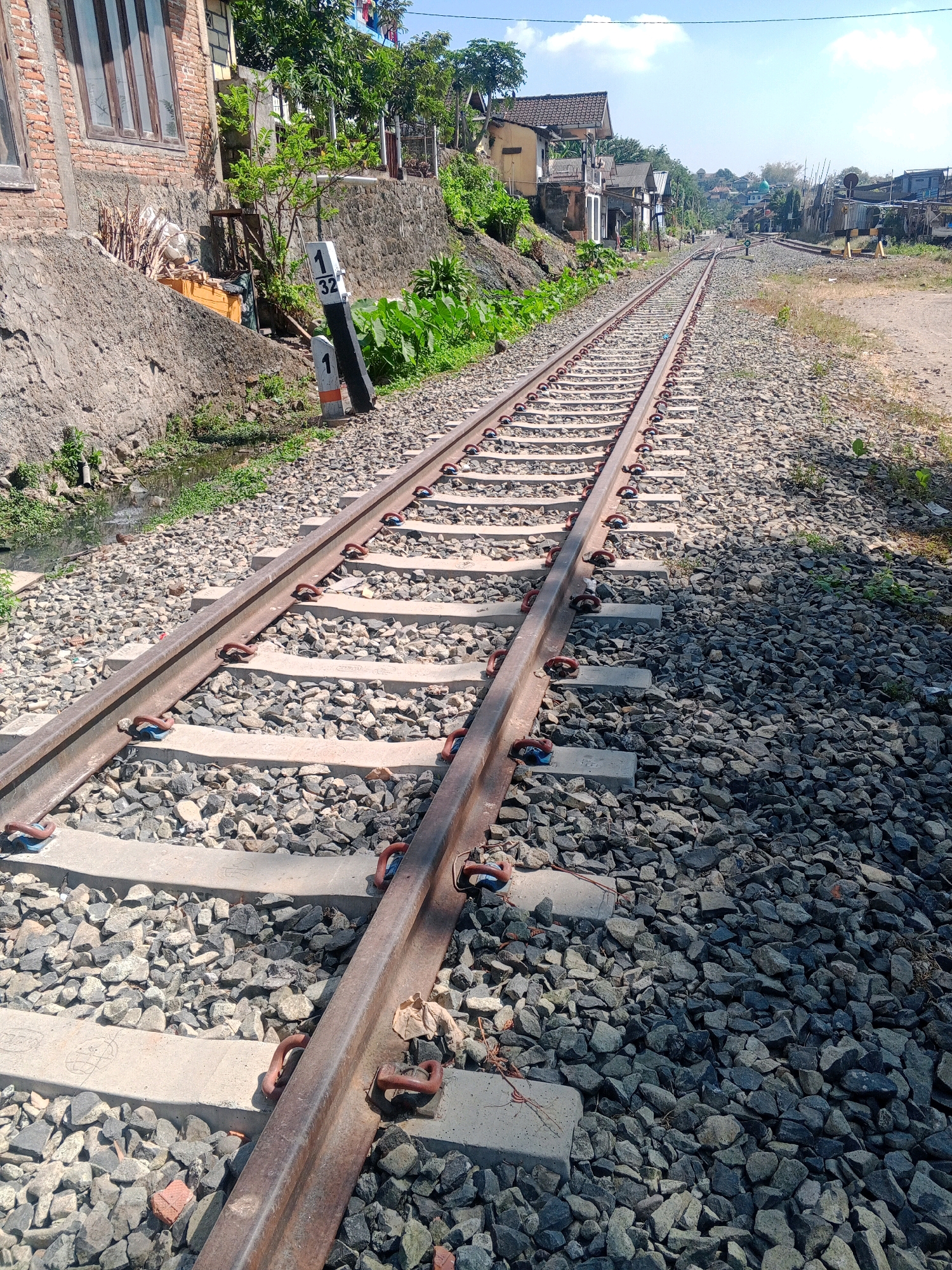 The image shows a view of a railway track in the city of Wonogiri, Indonesia. The track, made of steel rails on concrete sleepers, is surrounded by dark gray ballast. On the left side of the track, a low concrete wall borders a ditch where some greenery is growing. Beyond the wall, a brick building with large windows is visible, along with a pile of wood and a yellow object. A railway marker post with the numbers "1" and "32" is placed next to the track. The track stretches into the distance, where several houses and trees can be seen lining the sides. The sky is blue with scattered white clouds. The scene appears to be an outdoor, daytime setting.