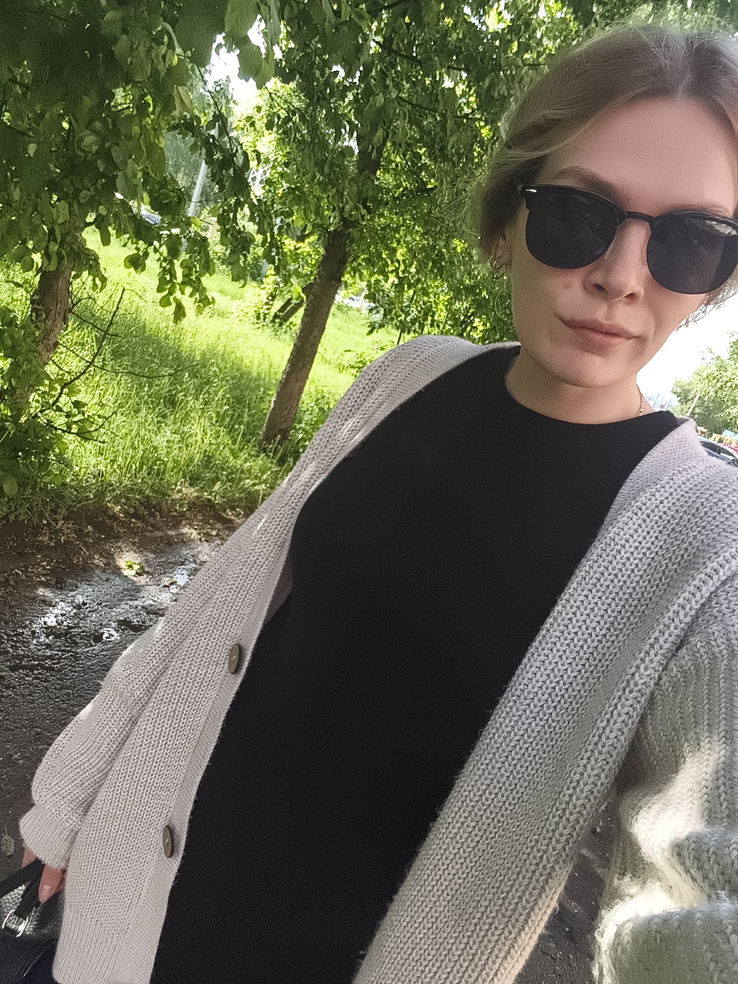 A woman wearing black sunglasses, a black top, and a light grey cardigan is standing outside on a path in Izhevsk, Russia. The path is wet and there are trees and green vegetation in the background.