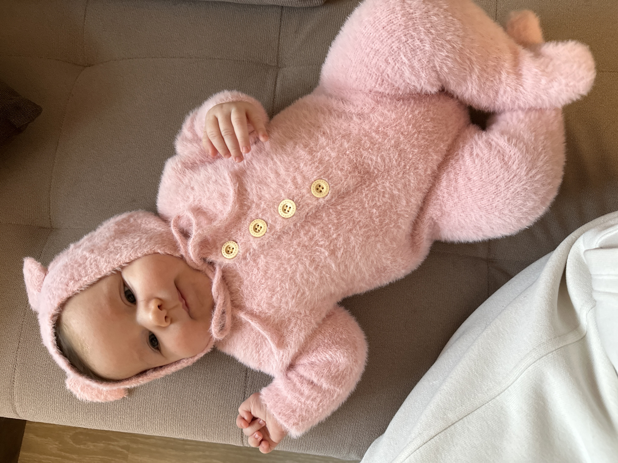 A baby wearing a pink, fuzzy bear-eared hooded jumpsuit with three tan buttons is lying on a tan upholstered surface. The baby's head is slightly tilted. A corner of a white garment is visible in the lower right corner of the image. The location is unknown.