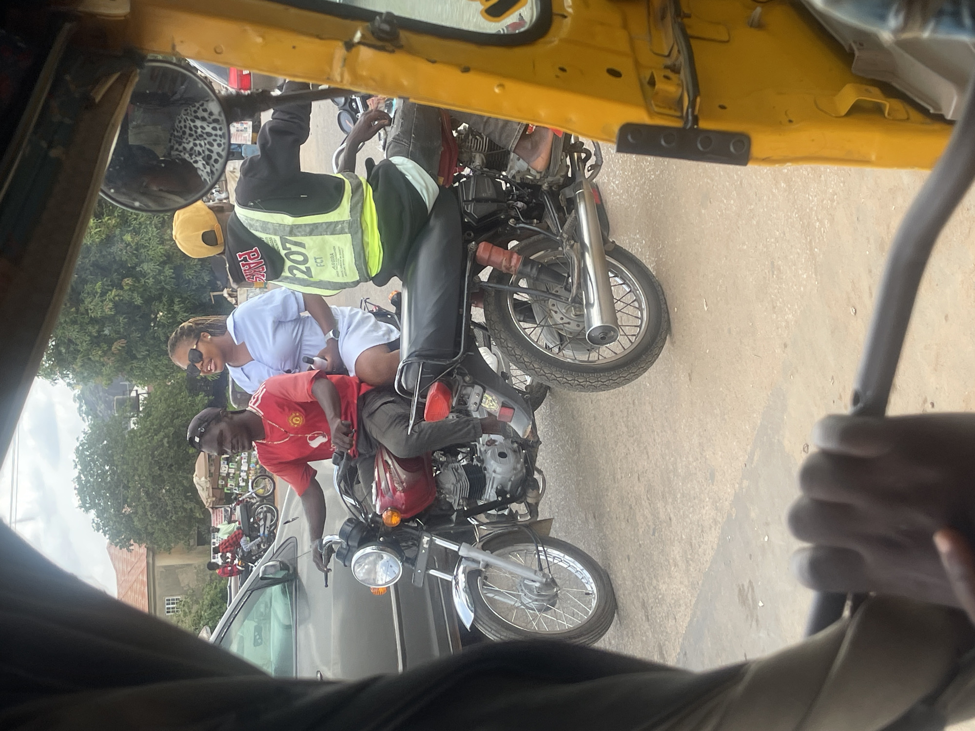 In Galadimawa Village, Nigeria, this image captures a street scene with multiple motorcycles and people. The foreground shows a blurred hand gripping a dark steering mechanism, likely from a yellow auto-rickshaw, with a portion of its yellow body visible on the right. In the middle ground, two motorcycles are prominent. The motorcycle on the left is red and driven by a man wearing a red t-shirt with a white logo and a black baseball cap. He is looking to his right. Behind him, on the same motorcycle, a woman wearing a light blue collared shirt and sunglasses is seated. To the right of this motorcycle, another motorcycle is visible, with a driver wearing a yellow baseball cap, a black hooded jacket over a lime green reflective vest labeled "207 FCT ARSRA," and dark pants. The background reveals trees, buildings with terracotta roofs, and a cloudy sky. A small stall with goods displayed on shelves is situated behind the motorcycles. The road surface appears to be asphalt or packed dirt.