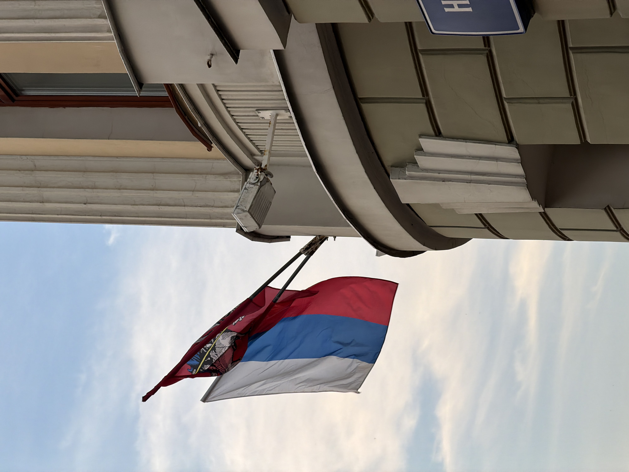 The image depicts a Russian flag and a red flag with a coat of arms flying from the side of a building in Moscow, Russia. The flags are attached to a metal pole extending from the building. The building appears to be made of light-colored stone with some architectural detailing. A street sign with the letter "H" in a blue square is visible on the right side of the image. The sky is cloudy.