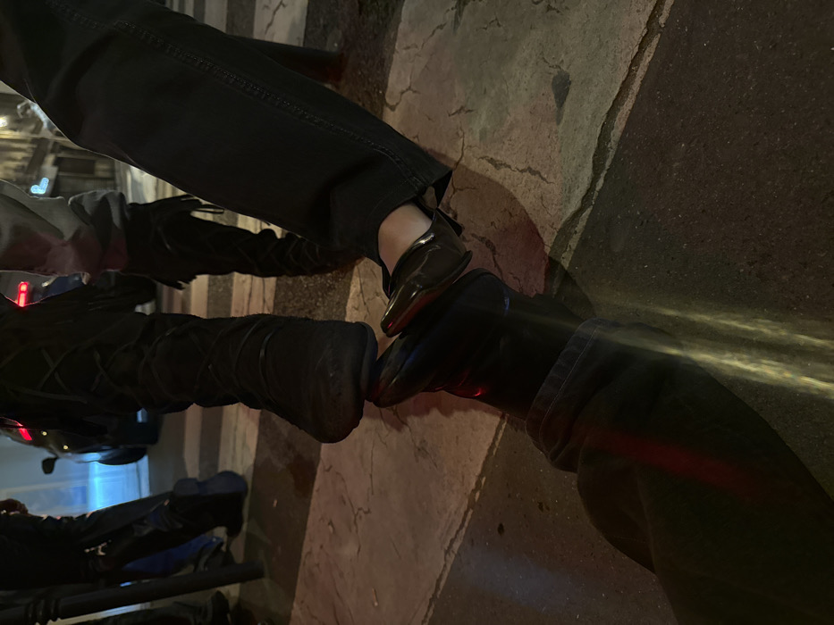 This low-angle photograph captures a detailed view of multiple individuals' lower legs and feet on an urban street at night in Paris, France. Artificial light sources illuminate the scene, casting shadows and reflections.
In the immediate foreground on the lower left, a person's left leg, dressed in dark pants and a dark, possibly leather, shoe or boot, is extended. A red light reflects prominently on the inner side of this shoe, and vertical yellowish light streaks appear on the pant leg.
Centrally positioned, a person wearing dark, knee-high boots with visible criss-cross lacing or decorative stitching has their left boot's toe resting directly on the left shoe of an adjacent person. This second individual, located to the mid-right, wears dark pants and dark, flat shoes featuring distinct red soles. The toe of this red-soled shoe is, in turn, resting upon the shoe of the person in the foreground. This arrangement signifies a direct, stacked interaction between the three sets of footwear.
In the blurred background, other individuals' lower legs and dark footwear are partially visible. A dark, vertical pole stands on the far left. A dark vehicle with illuminated red taillights is present in the deeper background, and a faint blue light source is discernible beyond it. The ground surface is composed of dark asphalt in the foreground, transitioning to a pedestrian crossing with white zebra stripes in the mid-ground. The distant background suggests multi-story stone or stucco buildings, characteristic of Parisian streetscapes.