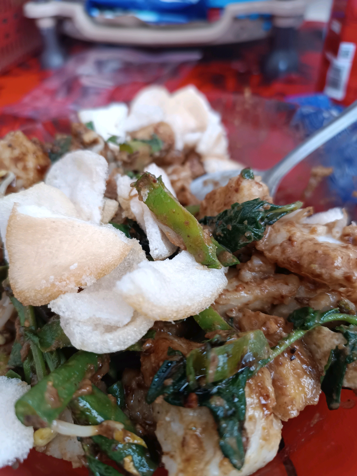A close-up, slightly overhead shot shows a bowl of Indonesian Gado-Gado salad, a local specialty from Tabanan, Indonesia. The salad is a colorful mix of vegetables, including long beans, bean sprouts, and leafy greens, all coated in a rich peanut sauce. Crispy crackers, known as 'krupuk', are scattered on top, adding a textural contrast. A fork is partially visible, suggesting the dish is ready to be eaten. The background is blurred, but a hint of a red tablecloth and a plastic container can be seen, indicating an outdoor setting, likely a street food stall or a casual dining spot.