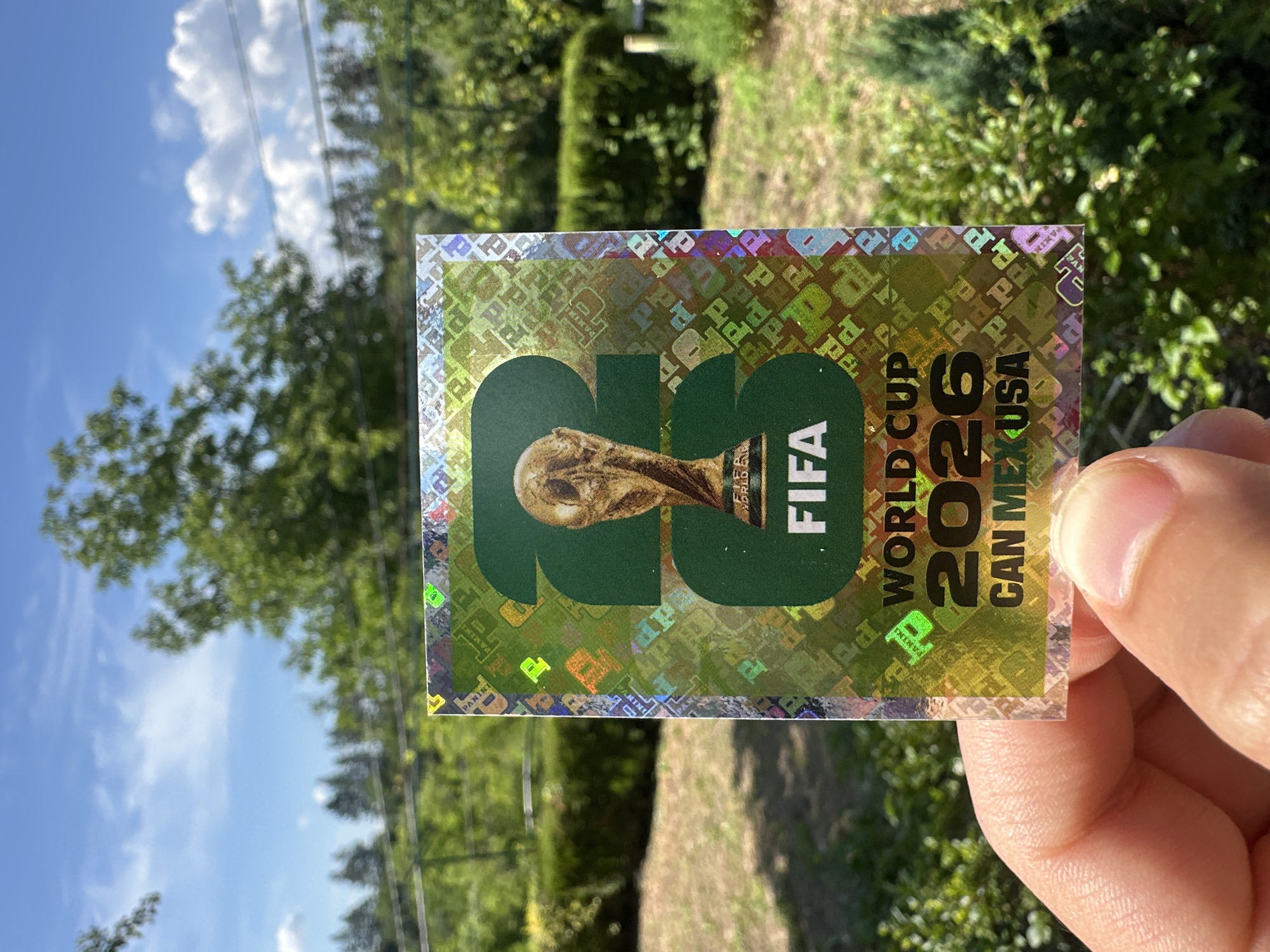 A hand is holding a rectangular sticker with a holographic background. The sticker features the FIFA World Cup 2026 logo, which includes the number "26" in large green digits and the FIFA logo. Below that, it reads "WORLD CUP" and "CAN MEX USA". The background of the sticker is a repeating pattern of the word "PANINI" in various colors and styles, interspersed with small FIFA logos. The sticker is shiny and catches the light, creating a shimmering effect. The background behind the sticker is blurred, showing greenery, trees, and a blue sky with clouds, suggesting an outdoor setting. The sunlight suggests it is daytime. The scene is likely in Prisovo, Bulgaria, given the location context.