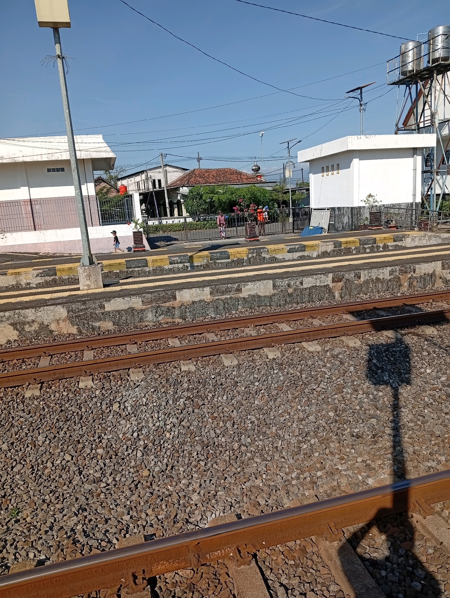 The image depicts a train station platform in Tebon, Indonesia, under a clear blue sky. In the foreground, two parallel train tracks are visible, filled with ballast. A shadow of a person holding a camera is cast across the right side of the tracks. Behind the tracks, a raised concrete platform runs along the length of the station. This platform is marked with yellow and black stripes, indicating a safety zone. To the left, a tall, light-colored pole with a rectangular fixture at the top stands near the platform. Beyond the platform, there are several buildings, including a white building with a flat roof and visible ventilation grilles, and other structures with tiled roofs. A fence separates the platform area from the buildings and surrounding greenery. A few people are visible on the platform, including a child running and a few adults standing or walking. Power lines are strung across the sky. In the background, more buildings and trees are visible, suggesting a residential or mixed-use area. The overall scene is one of an operational train station on a sunny day.