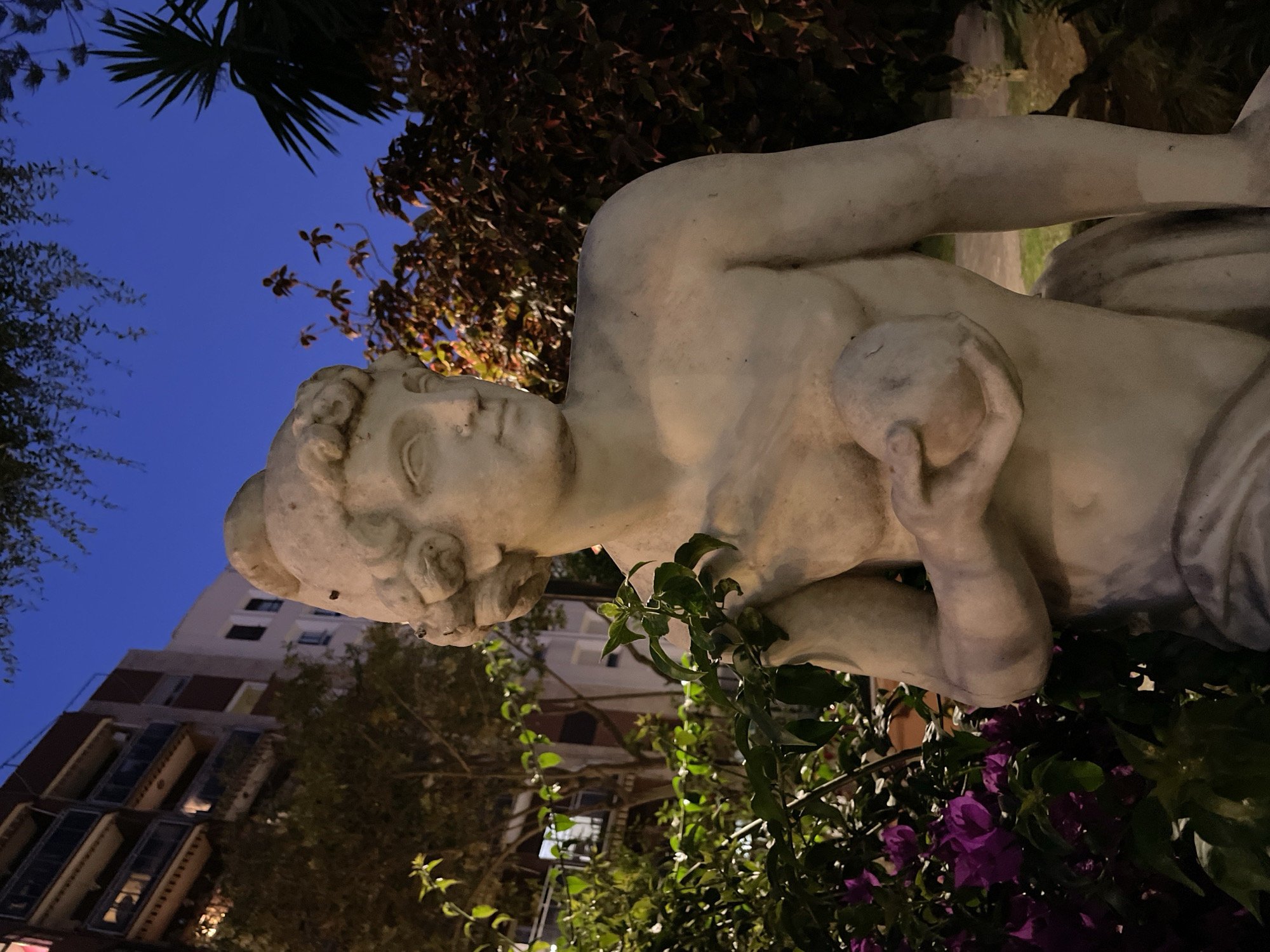 A stone statue of a young man holding a sphere is positioned in a garden at dusk. The statue is rendered in classical style, with idealized features and a muscular physique. He is wearing a helmet and a draped garment. The statue is surrounded by lush greenery, including flowering bushes with vibrant purple blossoms and leafy branches. In the background, a multi-story building with balconies and illuminated windows suggests an urban setting. The sky above is a deep blue, transitioning into twilight. The overall scene conveys a sense of serene beauty and classical artistry within a contemporary environment. The statue is likely located in La Línea de la Concepción, Spain, given the context of the query.