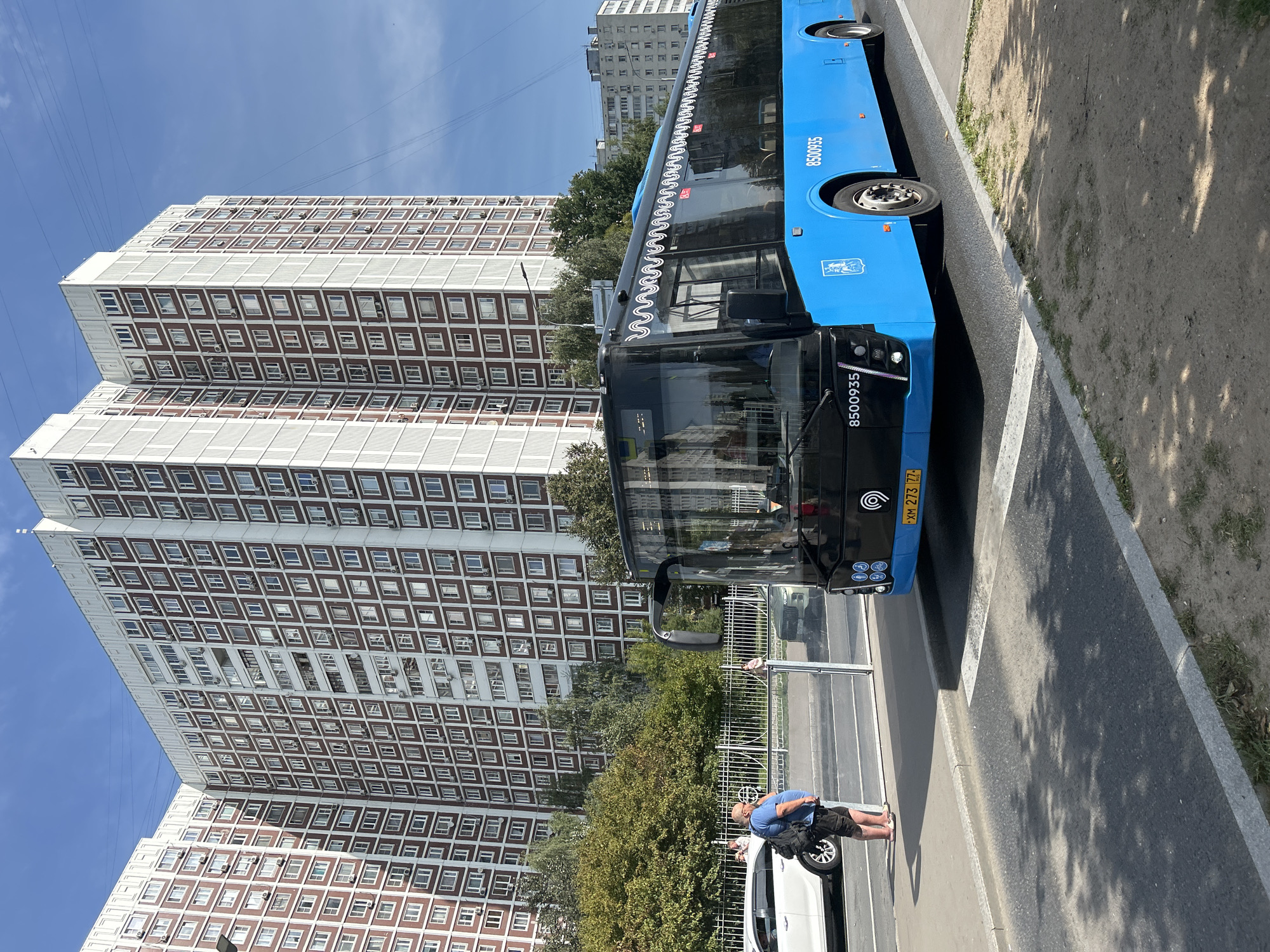 A bright blue public transport bus is stopped at a bus stop on a sunny day in Moscow, Russia. The bus, numbered 8500935, has its doors open and a few passengers are visible inside. In the foreground, a man with a backpack is standing on the sidewalk, facing away from the camera. He is wearing a blue t-shirt and black shorts. Behind the bus, there are tall, multi-story apartment buildings with many windows, characteristic of Moscow's residential architecture. Trees and greenery are also visible, suggesting a suburban or park-like setting within the city. The sky is a clear blue with a few wispy clouds. The license plate of the bus is visible and reads "XM 273 77 RUS".