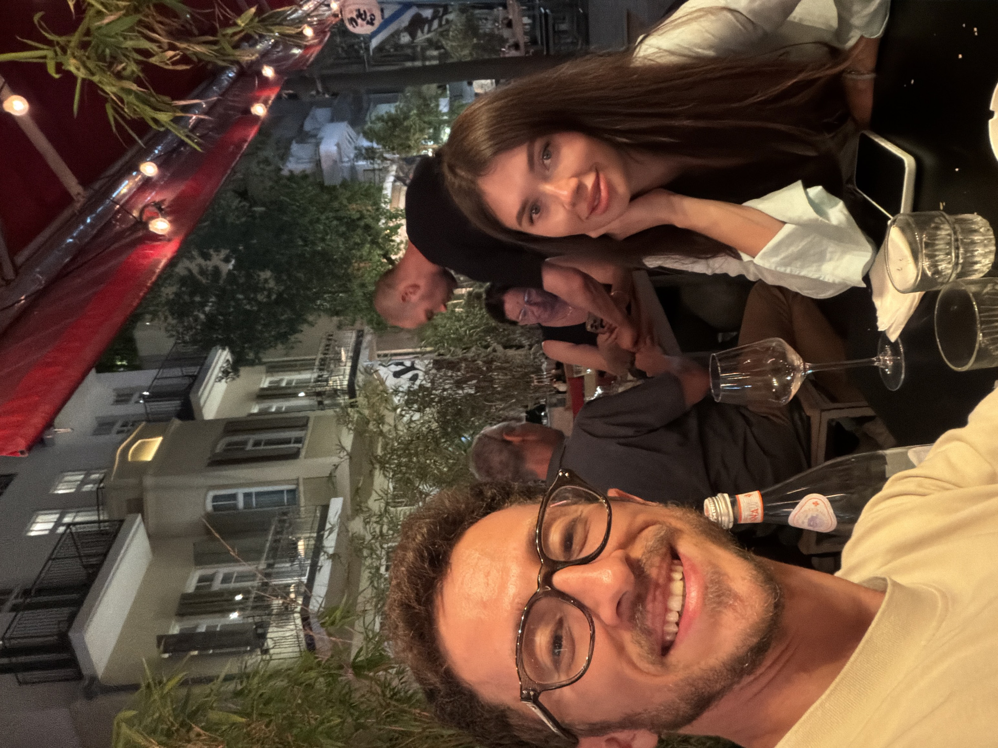 In a street-level outdoor dining area at night in Tel Aviv, Israel, four people are visible. In the foreground, a man with short, curly dark hair and glasses smiles at the camera, taking a selfie. To his right, a woman with long, dark hair rests her chin on her hand and looks at the camera. Behind them, a man wearing a black t-shirt is looking away from the camera, and a woman wearing glasses sits at a table. The environment includes bamboo plants, a building with lit windows and balconies, and a red awning with lights strung across it. On the table in front of the foreground subjects are two glasses, a bottle of San Pellegrino water, napkins, and a smartphone.