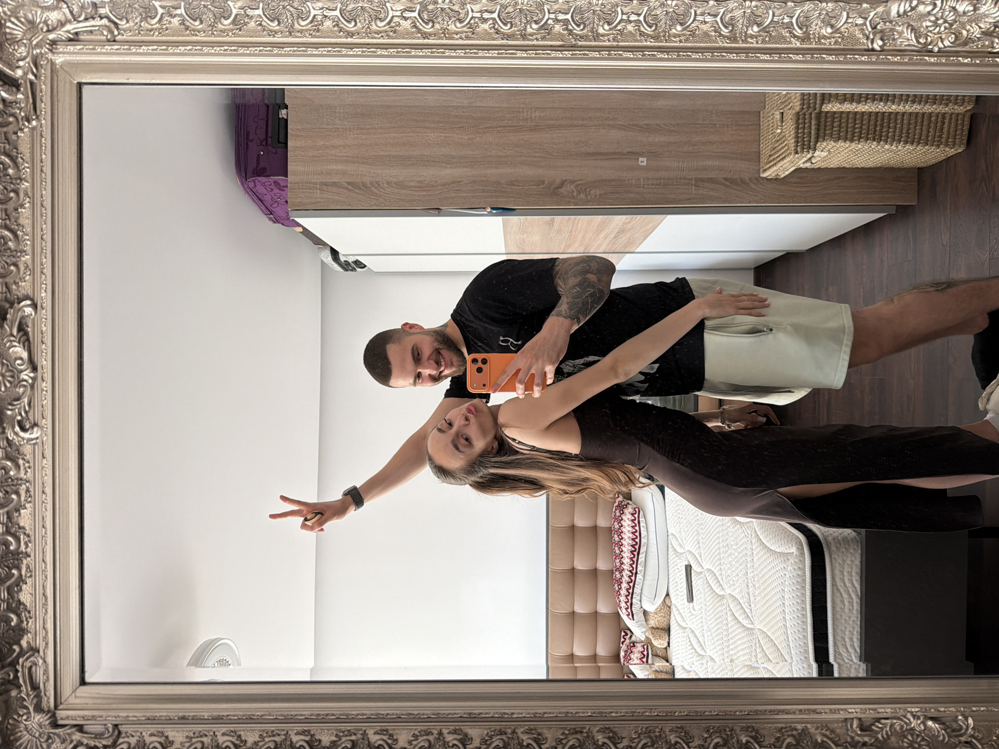 A couple is reflected in a framed mirror. The man has tattoos, a dark shirt and shorts. The woman is wearing a dress and is giving a peace sign. They are taking a selfie. Behind them is a bed, a closet, and a basket. The location is likely indoors, possibly in a bedroom. Sofia, Bulgaria is not identifiable in the image.