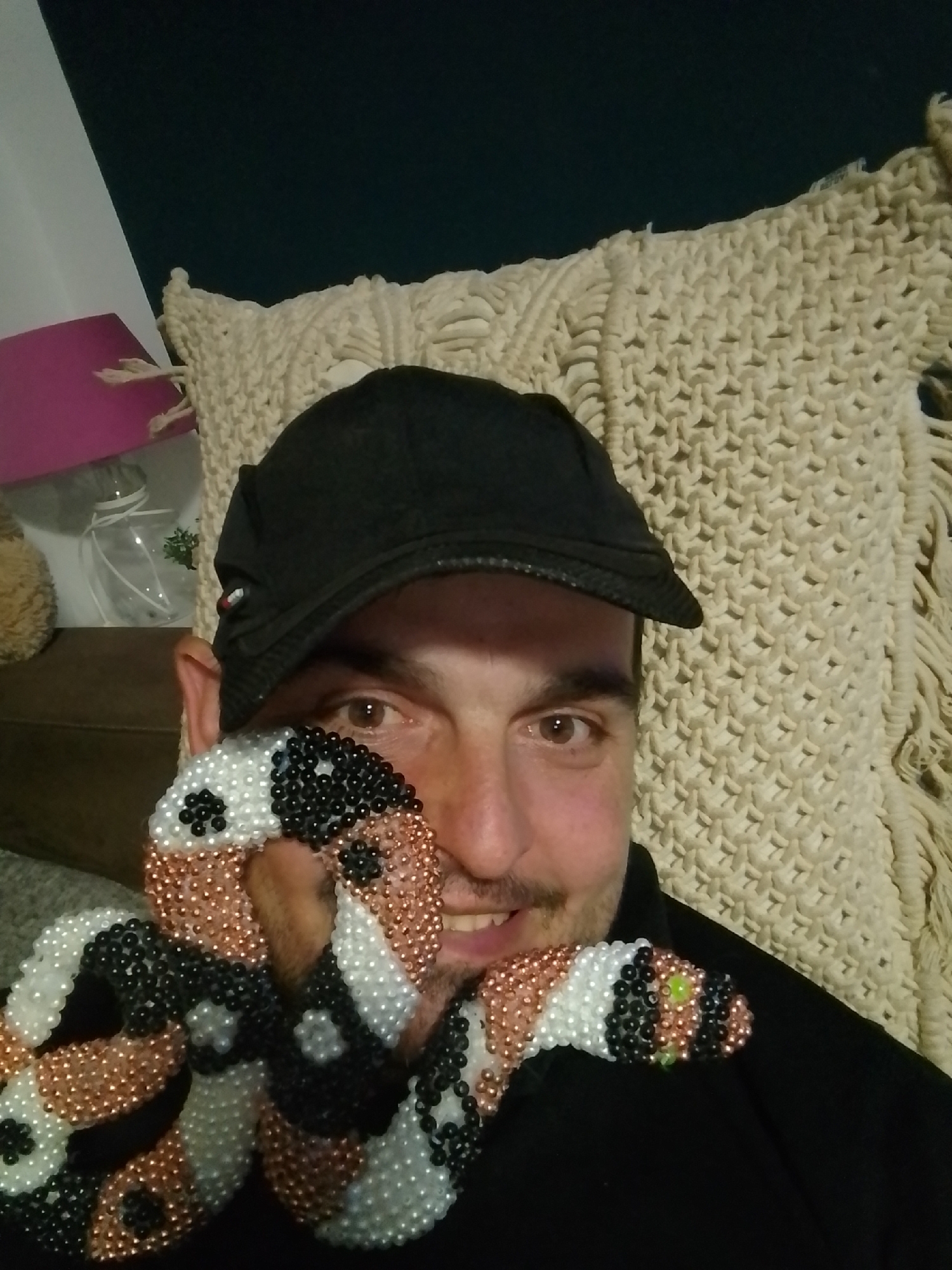 The image shows a person wearing a black baseball cap and a black shirt, holding a beaded snake figure in front of their face. The snake is multicolored, with white, black, and copper-colored beads. The person has brown eyes and a mustache, and is smiling. In the background, there is a textured cream-colored blanket or pillow, and a glimpse of a pink lampshade and a glass object. The lighting suggests an indoor setting. The location is not specified in the image.