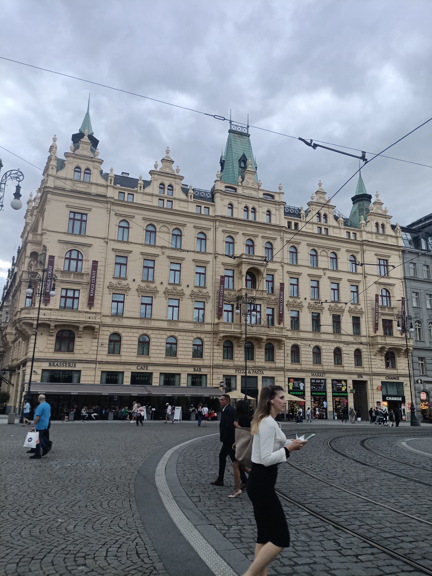This medium-shot, full-color photograph captures a street scene in Prague, Czechia. The central focus is a grand, ornate building with a pale yellow facade, adorned with multiple towers topped with green spires. The building features numerous windows and decorative architectural elements. Below, the ground floor houses various businesses, indicated by text labels such as "BRASSERIE," "CAFE," "BAR," "PIZZA & PASTA," "MARKET," "SPA," and "CHANGE." Banners with "KINGS COURT" are also visible.
The street is paved with cobblestones and features tram tracks. Several people are present, walking across the street or in the background. A woman in the foreground, wearing a white jacket and a black skirt, is walking while holding a piece of paper and wearing earbuds. A man in a blue shirt is walking away from the camera, carrying a shopping bag. Other individuals are seen in the background near the building's storefronts.
The sky is overcast with gray clouds, suggesting a cloudy or possibly rainy day. The lighting is diffused, indicating it's likely daytime, possibly late afternoon or early evening. The overall atmosphere appears to be that of a busy urban environment, with people going about their daily activities. The architectural style of the building and the presence of tram tracks are strong indicators of a European city, consistent with the provided location context of Prague.