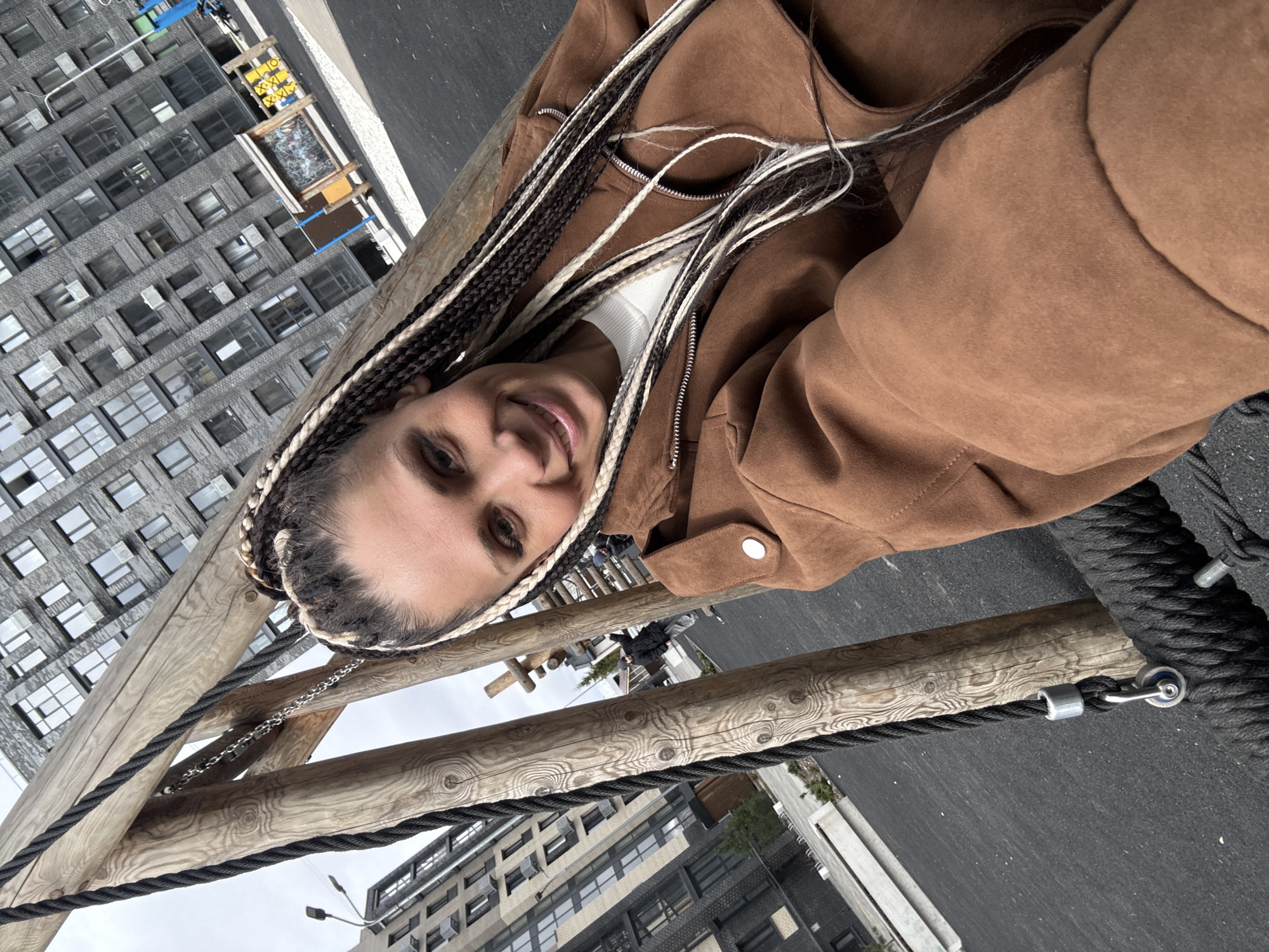 A woman is smiling in an outdoor photo, possibly a selfie, while standing on a wooden structure with thick black ropes. She has braids in her hair and is wearing a brown jacket. The background shows a modern multi-story building with numerous windows, indicative of an urban area. Based on the architecture and the presence of such modern structures, it could be in the city of Rusanovo, Russia.