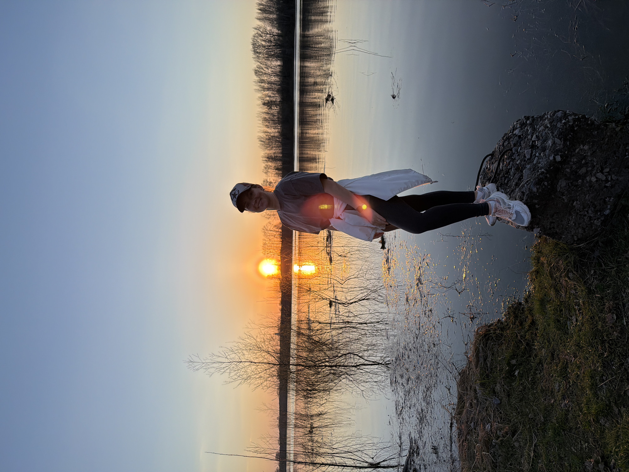 A person is sitting on a rock near a body of water, possibly a lake or a wide river. The sun is setting, casting an orange glow across the sky and reflecting on the water's surface. The setting sun is partially obscured by bare trees lining the opposite bank. The person is wearing dark leggings, a gray shirt, a white jacket, and athletic shoes. The location is likely Chaikovsky, Russia, considering the prompt's request. The rock the person sits on is located near the shoreline. The scene suggests a tranquil and picturesque evening.