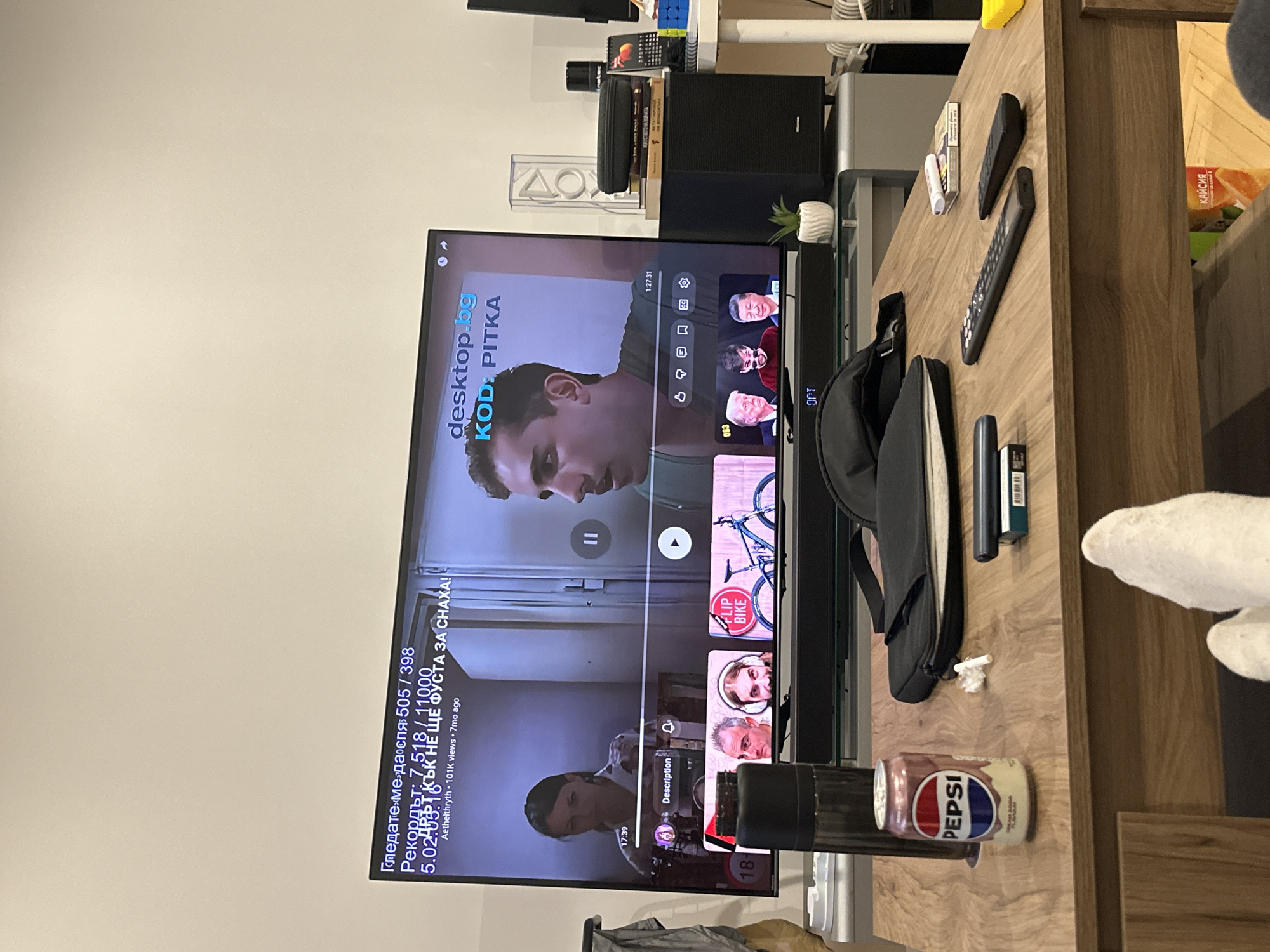 A medium, eye-level indoor shot shows a TV screen on a wooden surface, displaying a paused YouTube video. The video title appears to be "KOD: PITKA" from the desktop.bg channel. Text on the screen indicates a recording duration of "5.02:03:16" and reads "Гледате ме да спя 505/398 Рекордът: 7.518/11000 ДРФТ КЪК НЕ ЩЕ ФУСТА ЗА СНАХА!" A Pepsi can, a black bag, two remote controls, and a packet of cigarettes are on the wooden surface near the TV. In the top right corner, a container of "КАЙСИЯ" (apricots) is visible. A speaker is visible above the TV screen. The overall setting suggests a living room environment. There is no evidence to suggest a location in Lovech, Bulgaria.
