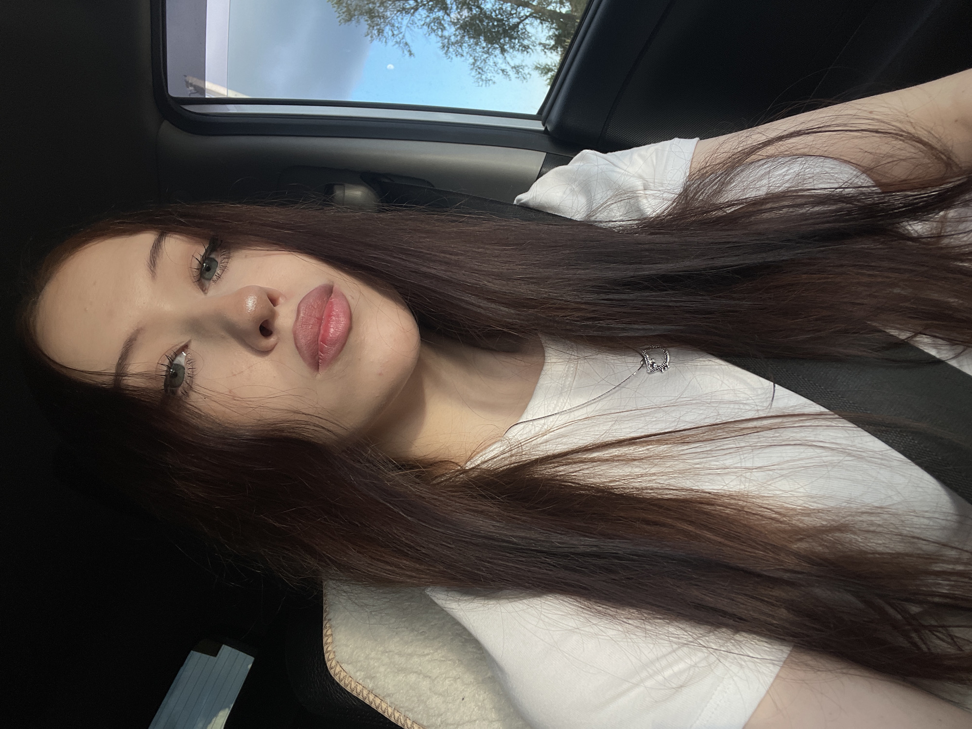 A young woman with long, dark brown hair is captured in a selfie from a slightly elevated angle, likely taken inside a vehicle. She is wearing a white t-shirt and a subtle necklace with a pendant. Her makeup is natural, highlighting her eyes and lips. The background shows the interior of a car, with a glimpse of the sky and trees through the sunroof, suggesting it's daytime. The lighting indicates it might be late afternoon or early evening. The overall mood is relaxed and casual. The image appears to be from Gavrilovka, Russia.