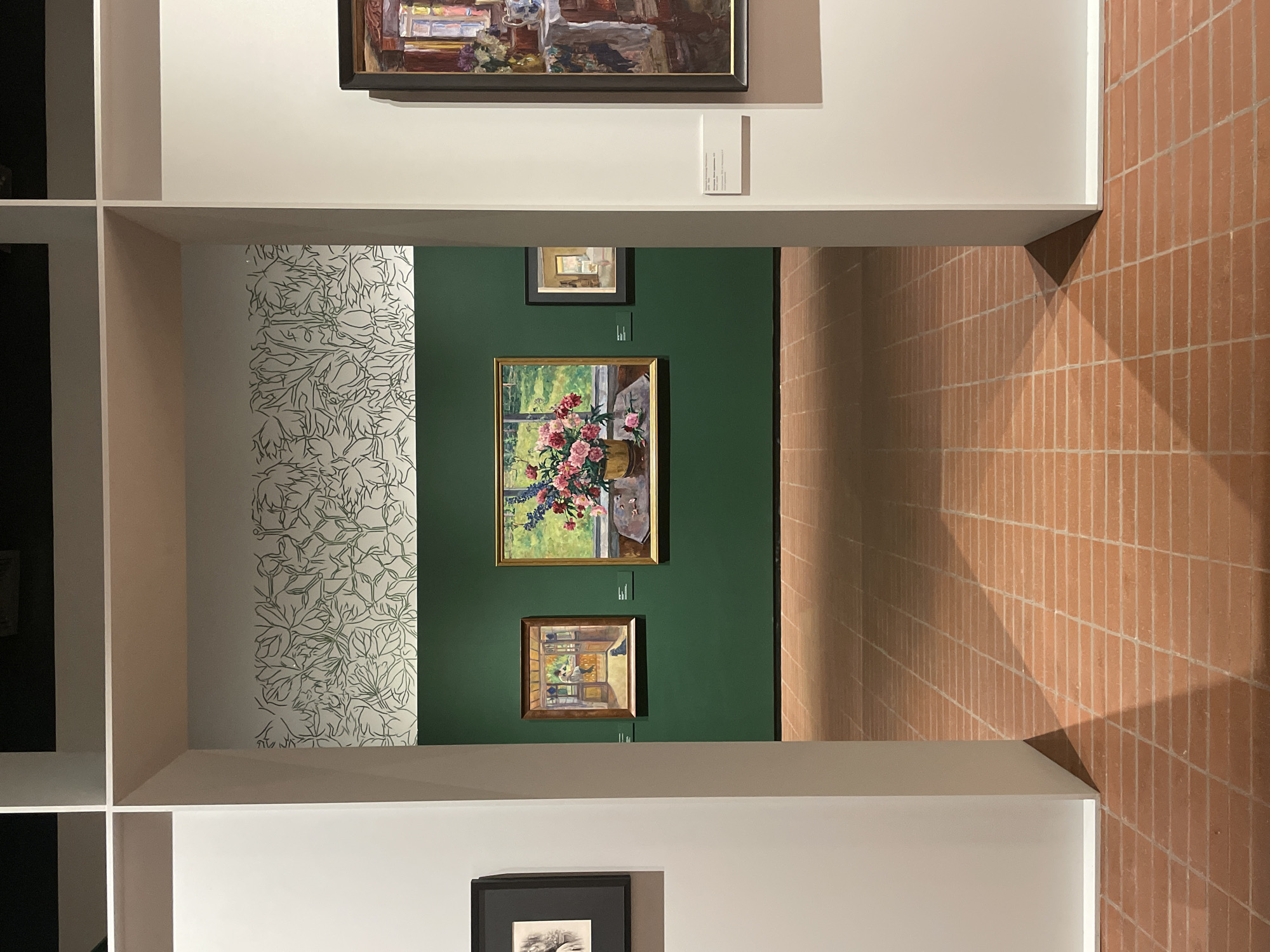 The image depicts an art gallery in Perm, Russia. The walls are painted in sections of white, green, and a patterned off-white. On the green wall are three framed paintings. The central painting is a floral still life in a golden frame. Above and below are two smaller paintings, also framed. The gallery is structured with a white shelving unit, framing the view of the paintings. To the right, a wall made of small, reddish-brown tiles is visible. Above the gallery view, another framed painting is partially visible. Below, a black and white framed picture sits on a shelf.
