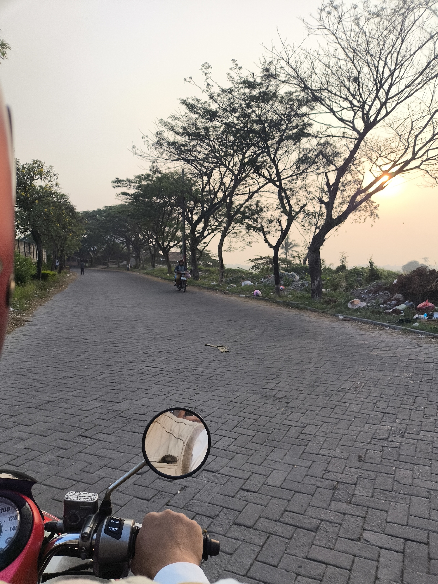 The image depicts a paved road lined with trees on both sides. The road is constructed with grey interlocking bricks. On the right side of the road, there is a significant accumulation of garbage. In the foreground, a person's hand is visible on the handlebars of a red motorcycle, with a rearview mirror reflecting a beige jacket. A speedometer with markings up to 140 is also visible on the motorcycle. In the distance, there are a few people walking and a person riding a motorcycle. The sun is setting, casting a warm glow over the scene, and its rays are visible through the branches of a bare tree. The sky is a pale orange. The location is identified as Cilegon, Indonesia.