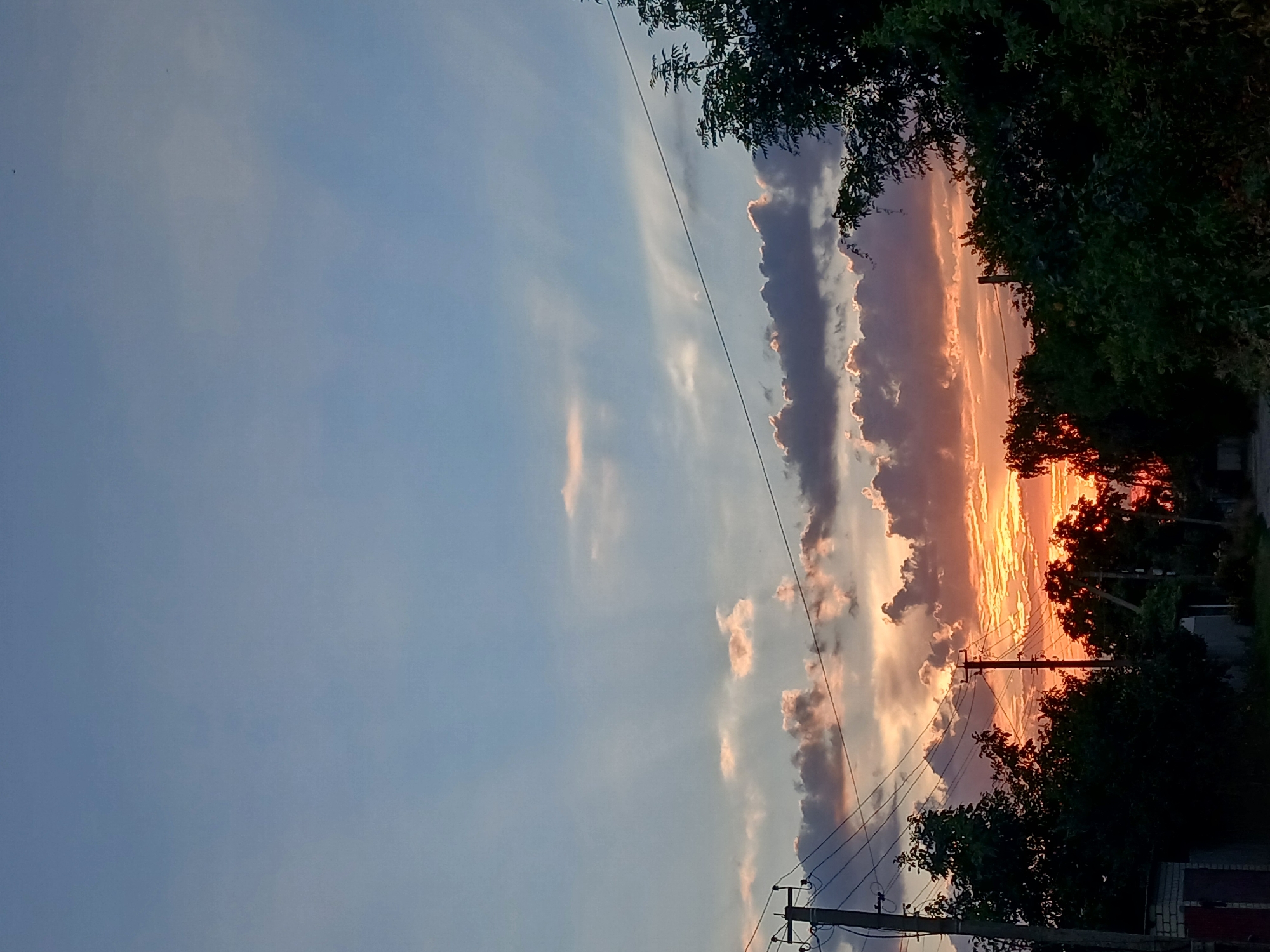 A low-angle, outdoor, eye-level shot depicts a rural road at sunset. The sky is a mixture of blues, grays, and oranges, with clouds tinged pink and yellow by the setting sun. Electrical poles and wires crisscross the scene, silhouetted against the vibrant sky. Dense green trees line the road, their foliage dark and lush. The foreground shows the edge of a building with a reddish-brown door. The overall impression is of a peaceful, slightly overcast evening in a rural setting.