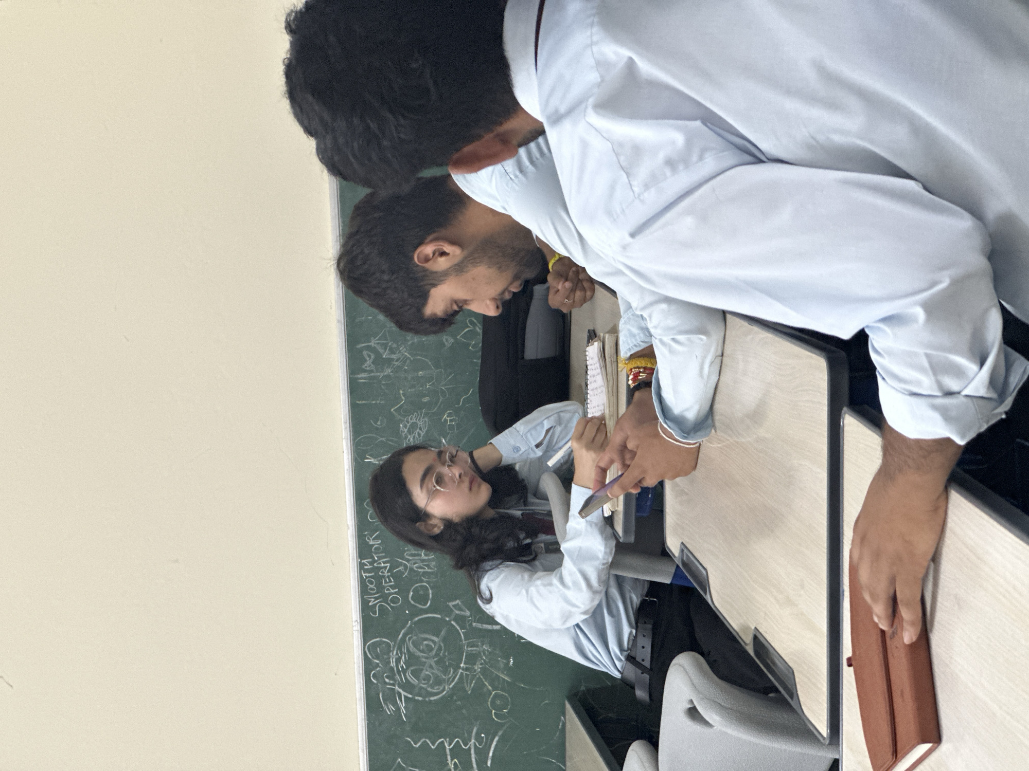In an indoor scene, likely a classroom, three young individuals are seated at desks. A young woman in the foreground, wearing a light blue collared shirt and glasses, is focused on writing in a notebook while holding a pen in her right hand. Her left hand is resting on her cheek, and she is looking down at her notebook. A mobile phone is visible in her hand.
Behind her, a young man with a beard, also wearing a light blue collared shirt, is looking towards his left, his hands clasped in front of him. To the far right, the back of another young person's head and shoulders are visible, also wearing a light blue collared shirt. They appear to be looking towards the left.
A green chalkboard is visible in the background with handwritten text and drawings, including the words "SMOOTH OPERATOR" and some cartoonish figures. The desks are made of wood with metal frames. The lighting suggests an overhead light source, casting some shadows. The overall atmosphere appears to be one of study or a casual classroom interaction.