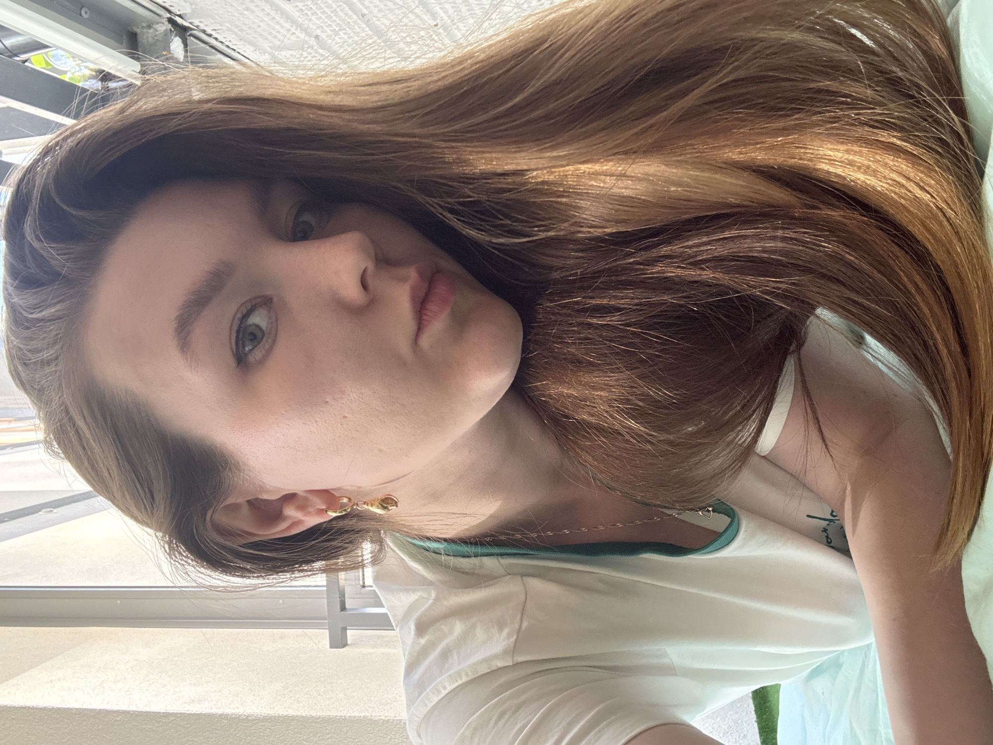 A young woman with long, light brown hair is shown in a close-up, selfie-style shot. She has fair skin, light eyes, and is wearing a white t-shirt with a green neckline. She is wearing gold earrings and a delicate gold necklace. The background features a window with a metal frame and a light-colored wall. There is no evidence of Izhevsk, Russia in the image.
