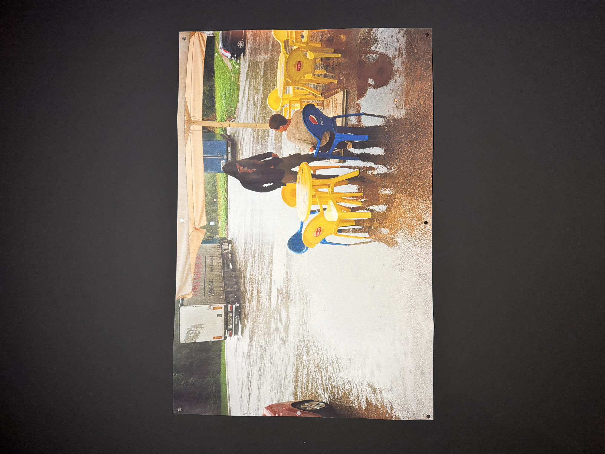 The image captures a flooded outdoor scene, likely a roadside cafe or rest stop. The ground is submerged under a significant layer of water, reflecting the sky and objects around.
Two individuals are visible amidst the flood. One person, wearing a dark jacket, stands in the midground. Another person, dressed in a light-colored top, is seated on a blue plastic chair that features the "Pepsi" logo. Numerous other plastic chairs, predominantly yellow and bearing the "Lipton" logo, and matching yellow plastic tables are scattered across the water-covered area. A large beige or light brown canopy structure provides overhead cover for a section of the seating area.
In the background, a large white truck trailer is parked on the flooded surface. Text visible on its side includes "www.scann.ro" along with other illegible writing. A blue shipping container is also present behind the people. In the foreground, a dark-colored car, possibly red or maroon, is partially visible, with its wheels submerged in the floodwaters. Another dark car can be seen further in the background.
The scene appears to be set during the daytime, likely under overcast skies or after heavy rainfall, given the extensive flooding. The presence of commercial beverage branding and large vehicles suggests a location adjacent to a road or highway. The context provided indicates this event took place in Moscow, Russia.
